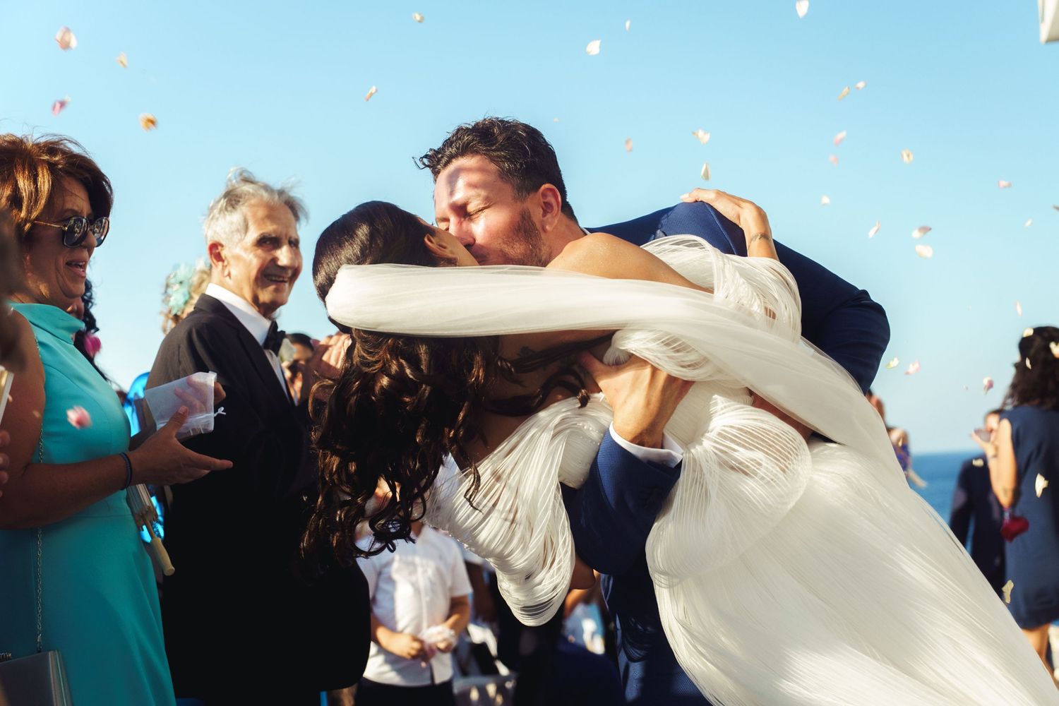 Wedding celebration with guests throwing confetti and celebrating outdoors on a sunny day.