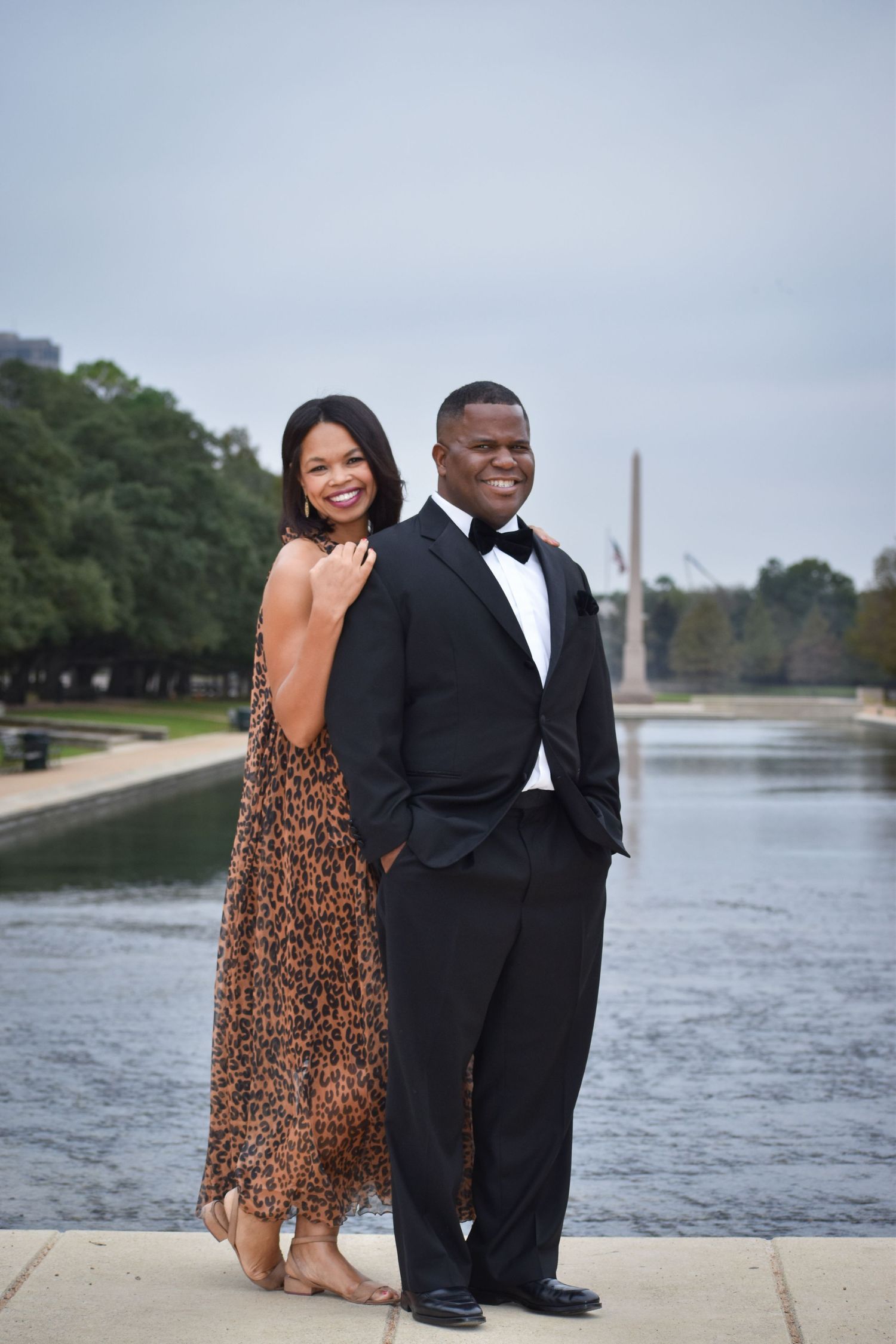 Elegant couple in formal attire posing by a reflective pool with city skyline in the background.