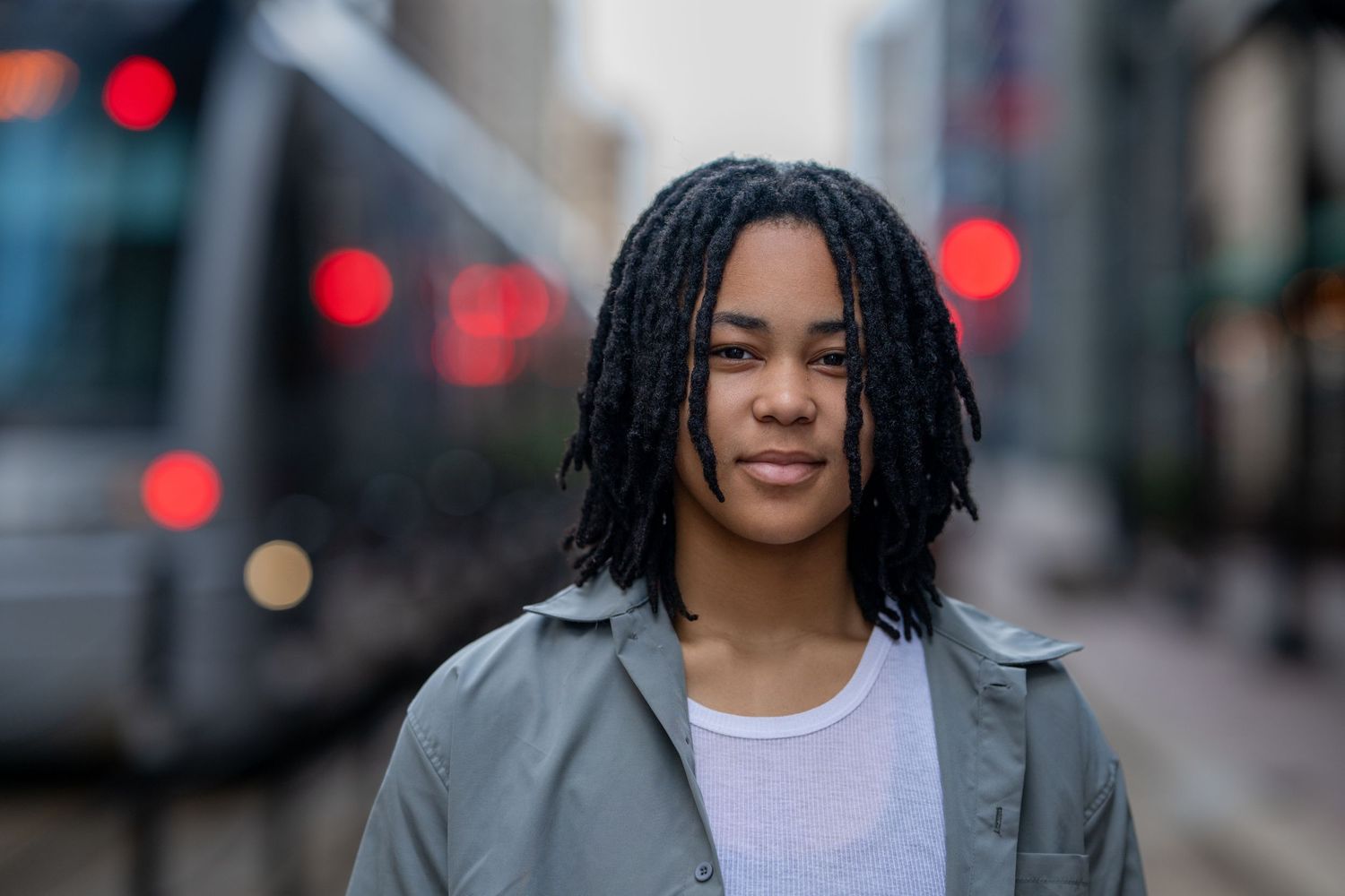 Young Black man with locs wearing a grey jacket poses on a city street with bokeh lights.