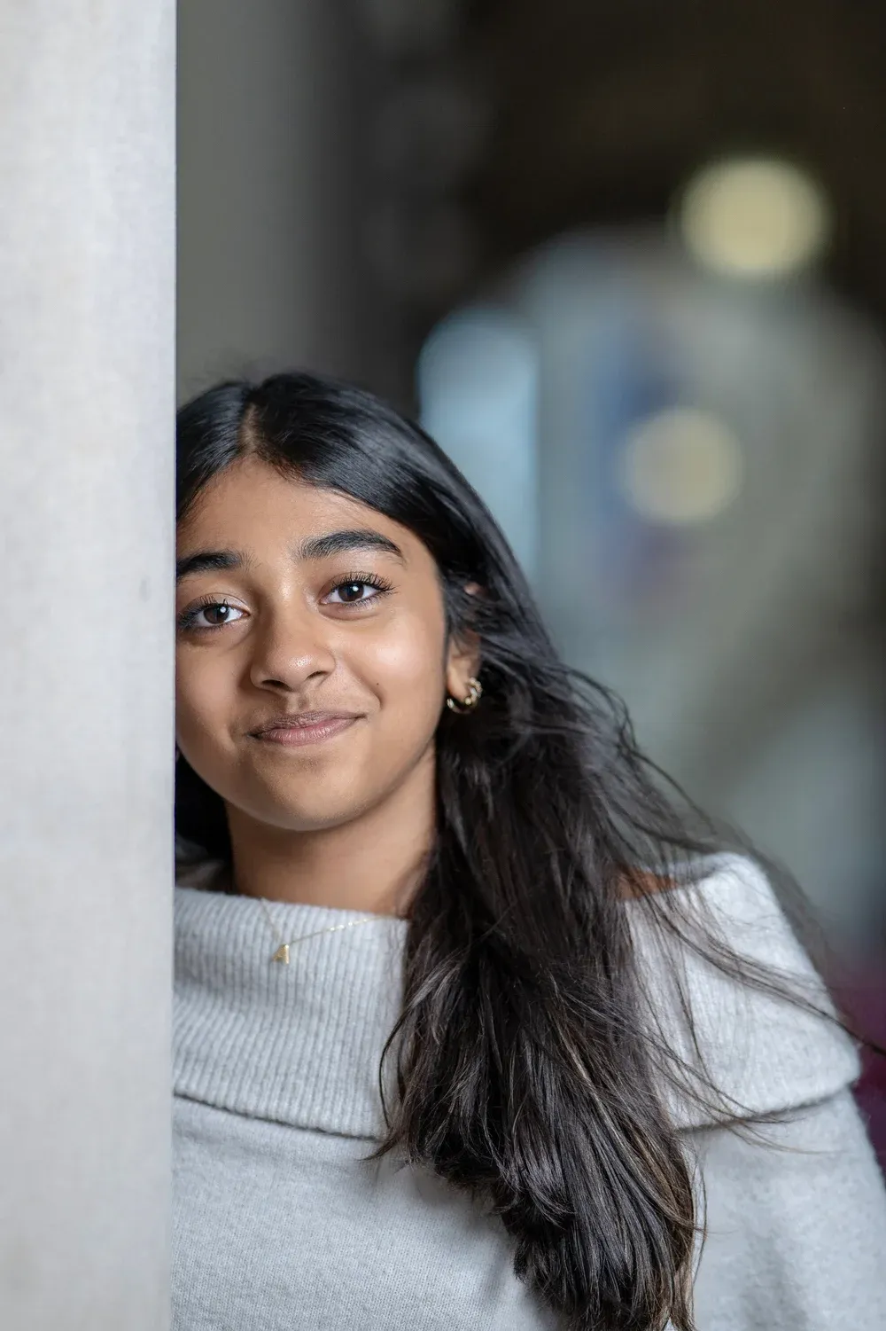 Young Indian girl with long black hair leaning against a white pillar, smiling softly.