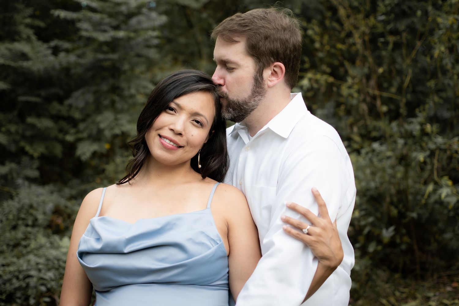 Romantic couple embracing outdoors, man kissing woman's cheek as she smiles in a light blue dress.