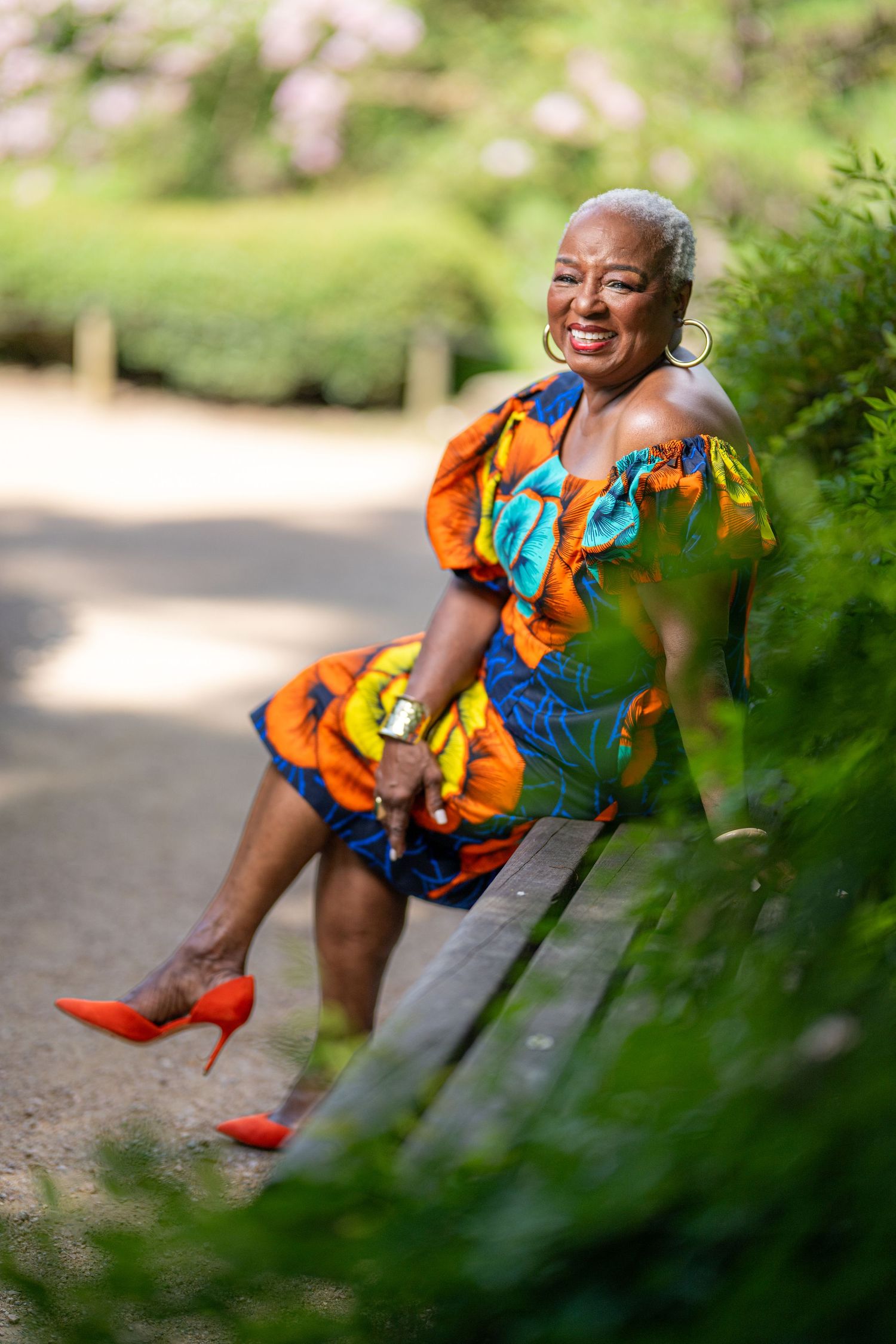 Elegant older woman in colorful floral dress and red heels sitting on park bench, smiling joyfully outdoors.