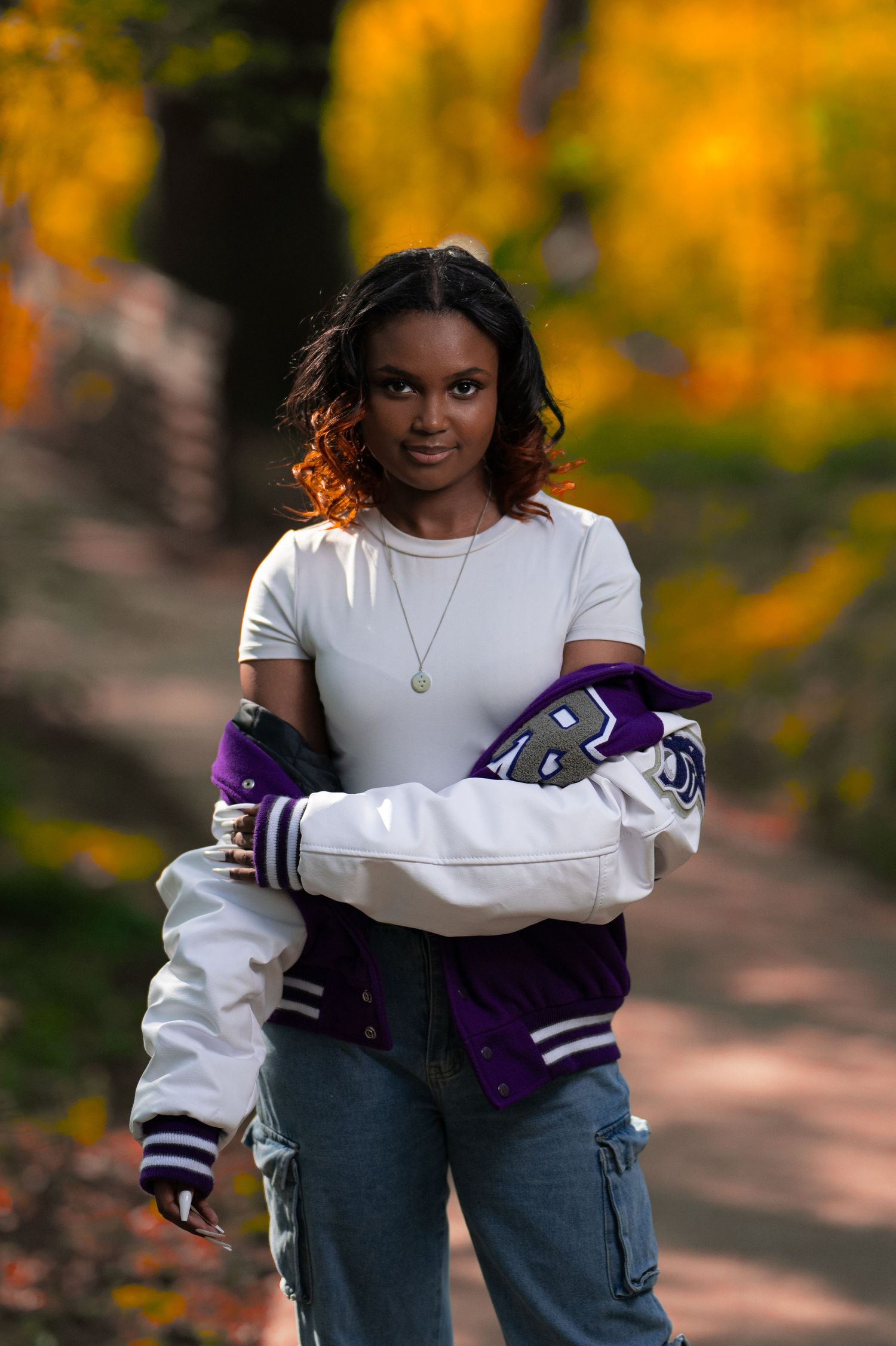 A young woman in a white tee and purple varsity jacket poses on a tree-lined autumn path.