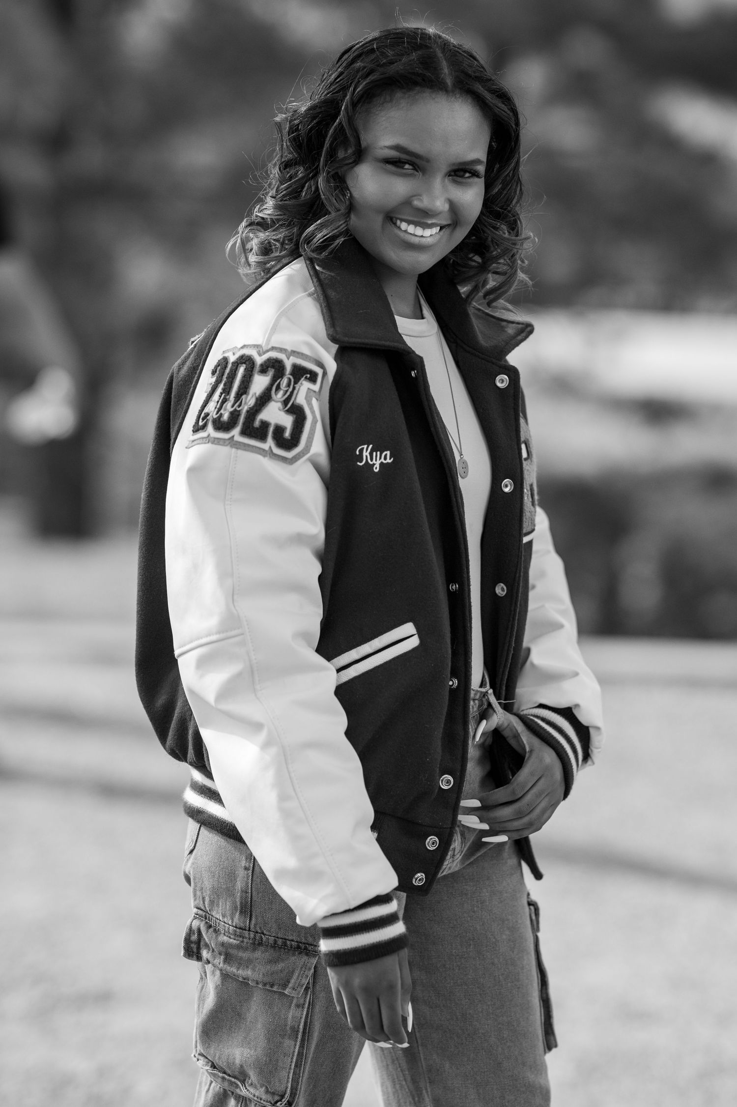 A smiling young woman in a 2025 varsity jacket poses outdoors in a black and white portrait photo.