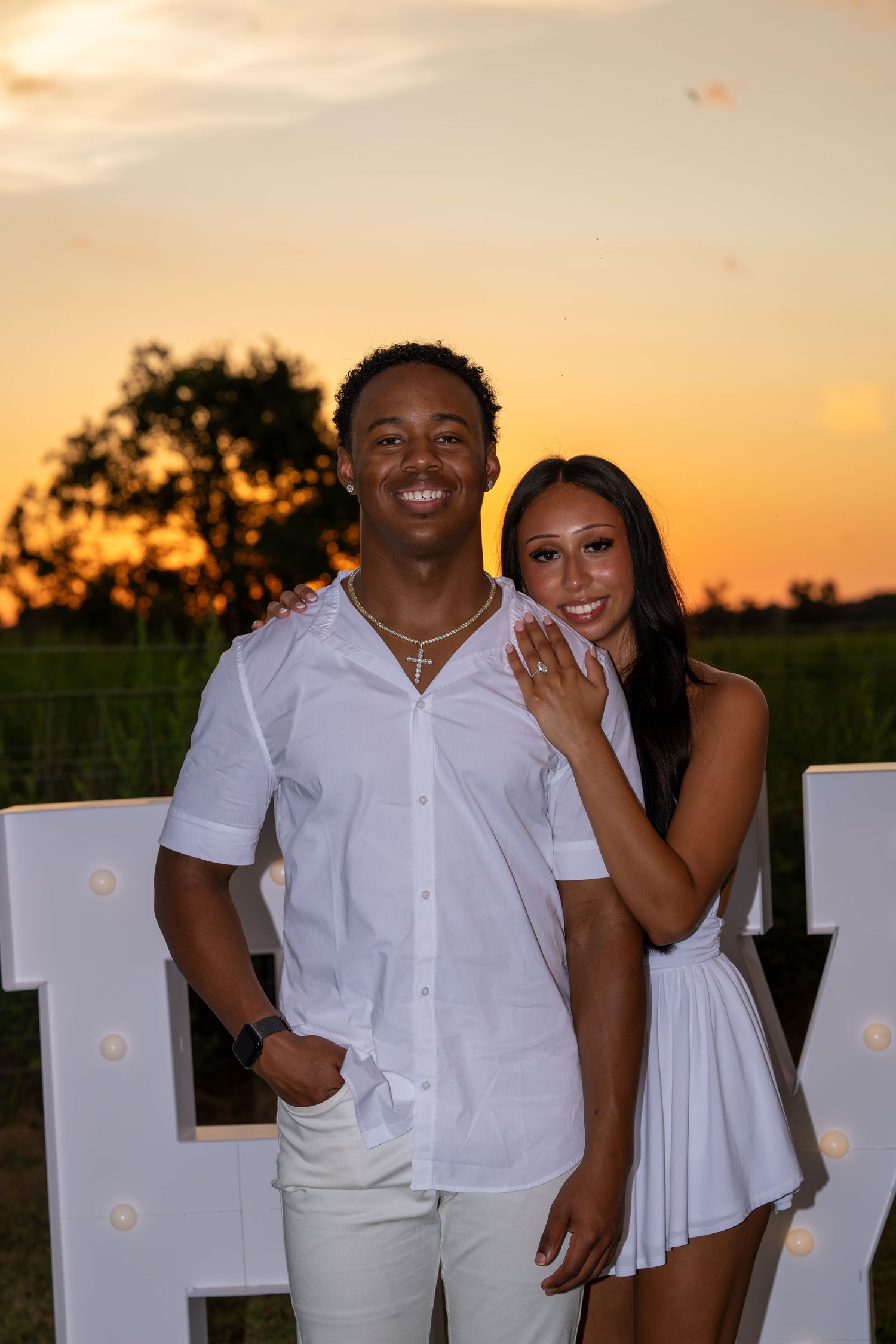 A smiling young couple in matching white outfits poses together at sunset near illuminated marquee letters.