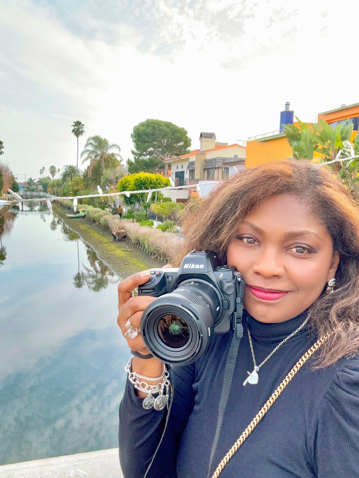 Woman holding a Nikon camera smiling in a selfie by a scenic canal with colorful buildings.
