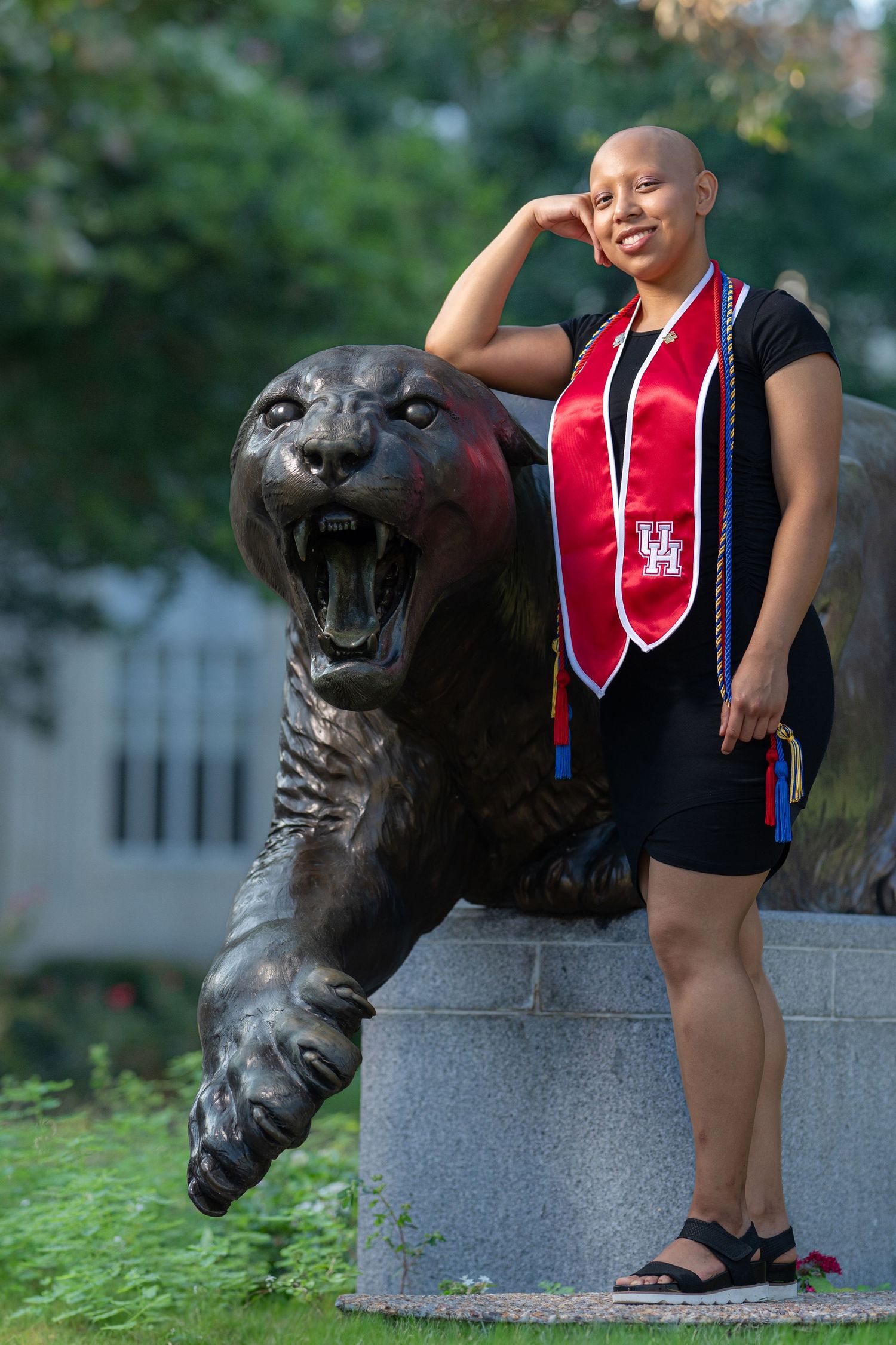 Graduate in black gown with red honor stole posing beside a cougar statue on campus.