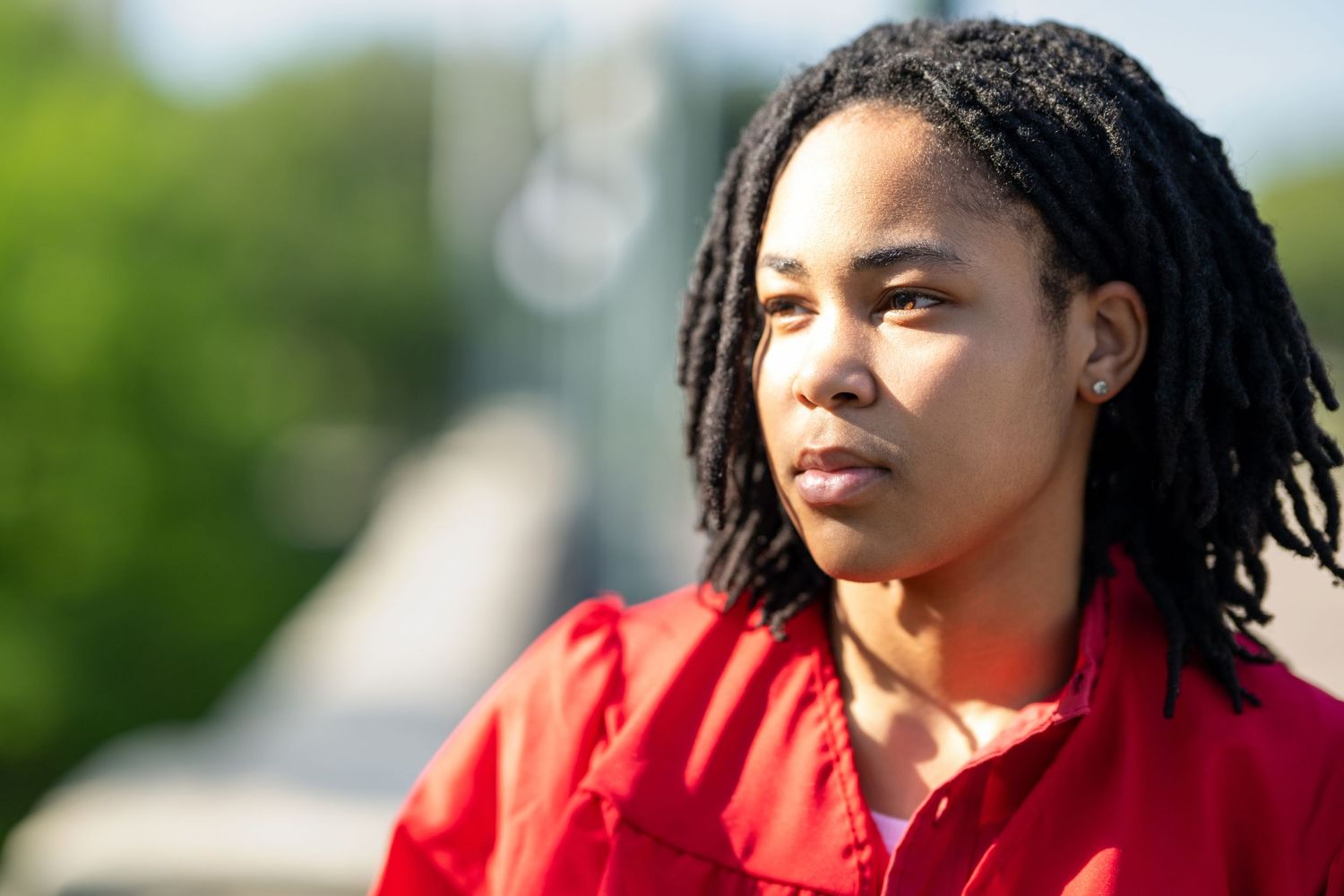 Young Black woman with locs wearing a red jacket gazing thoughtfully outdoors in sunlight.