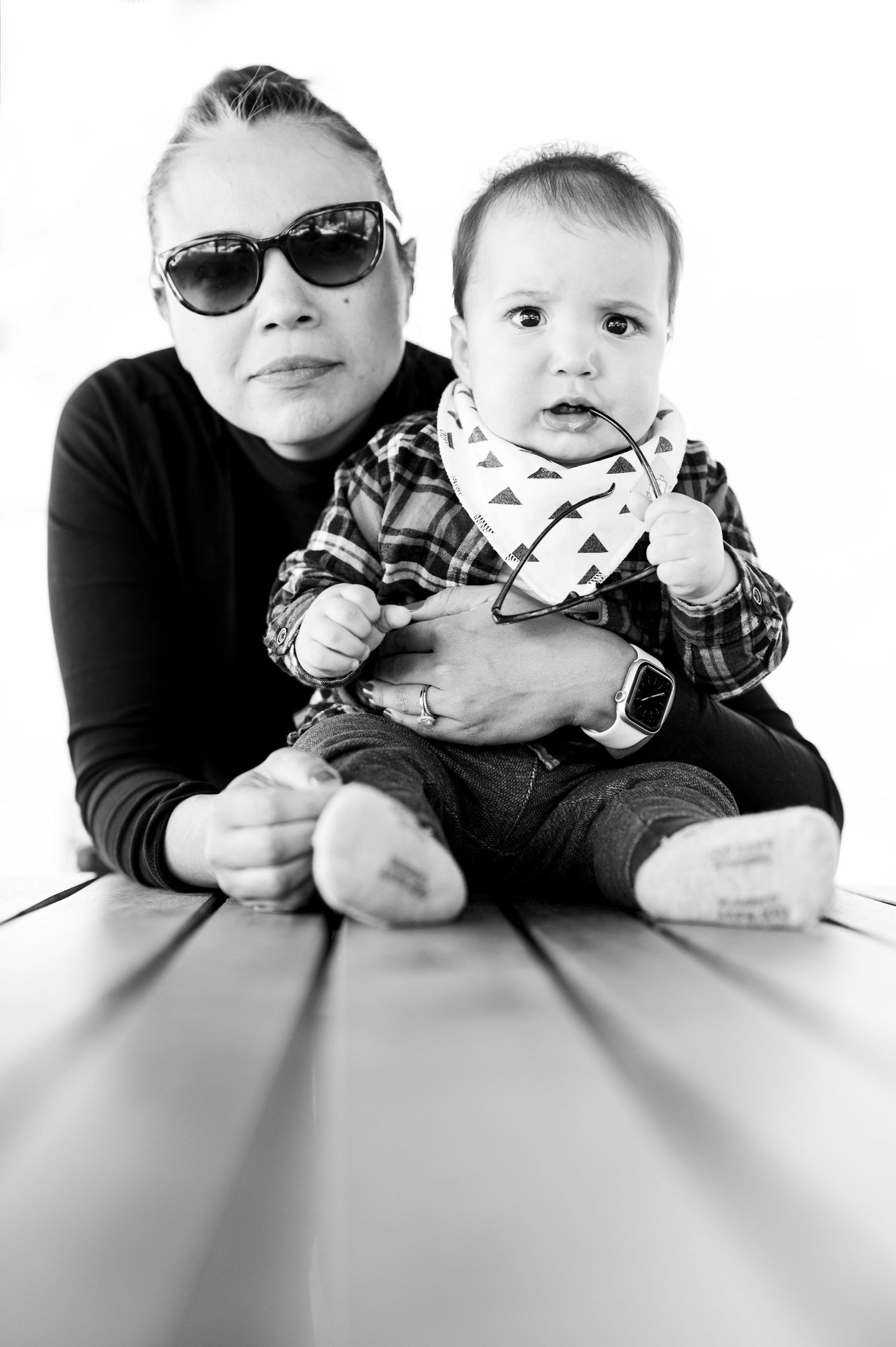 Black and white portrait of woman with sunglasses holding curious baby on table.