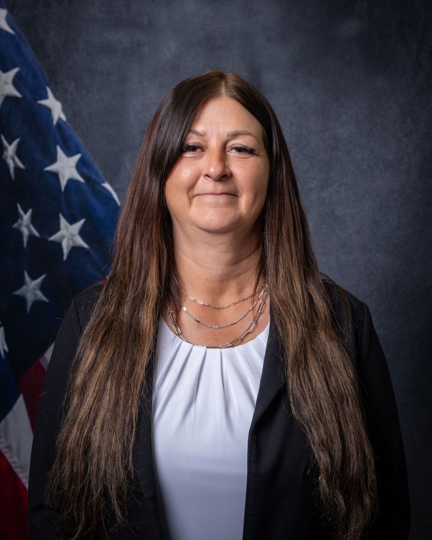 Official portrait of a woman with long dark hair wearing a black blazer and white top before an American flag.