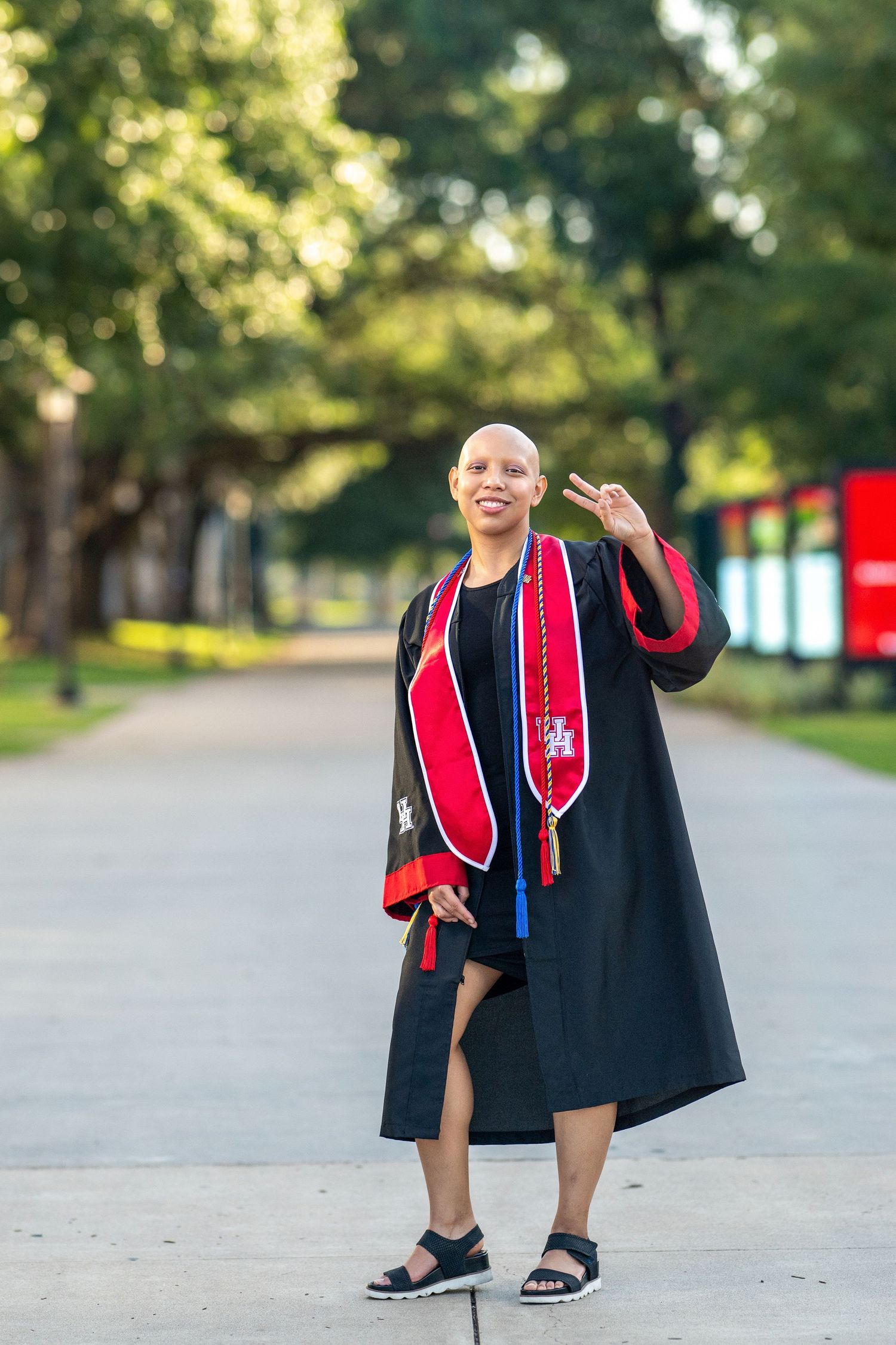 Smiling bald woman in graduation gown with red stole giving peace sign on campus path.