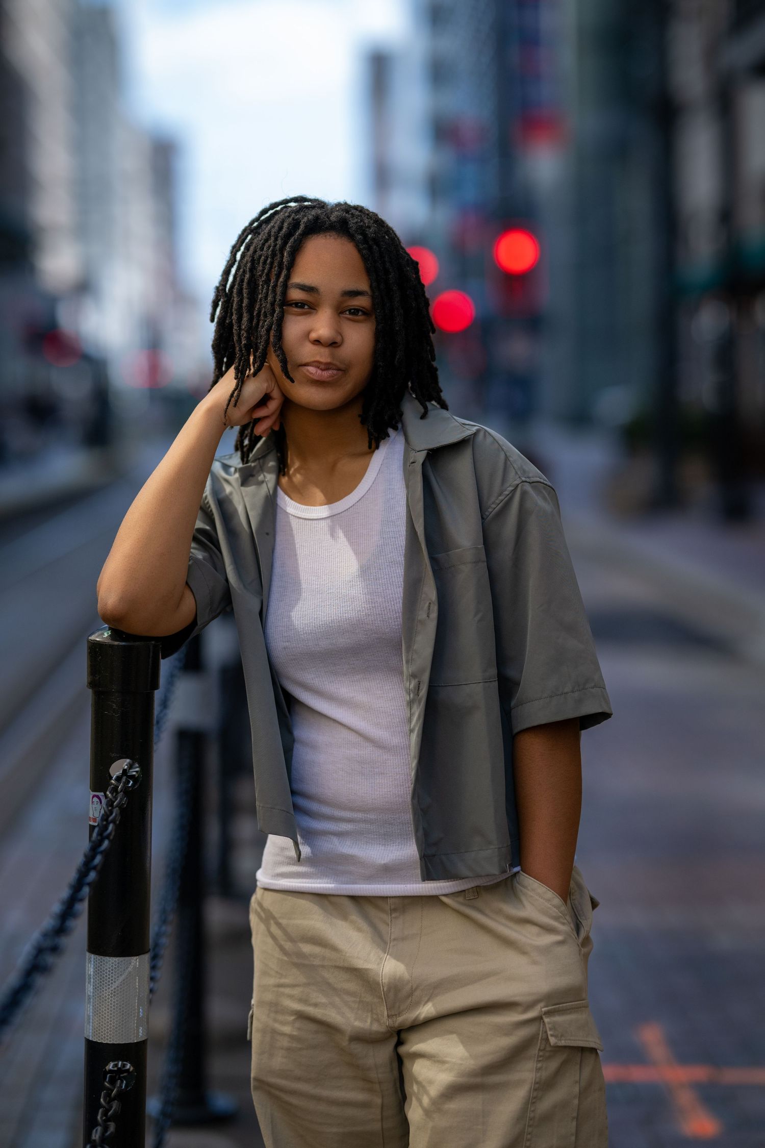 Young woman with locs in gray shirt and khaki pants leaning on a pole on city street.
