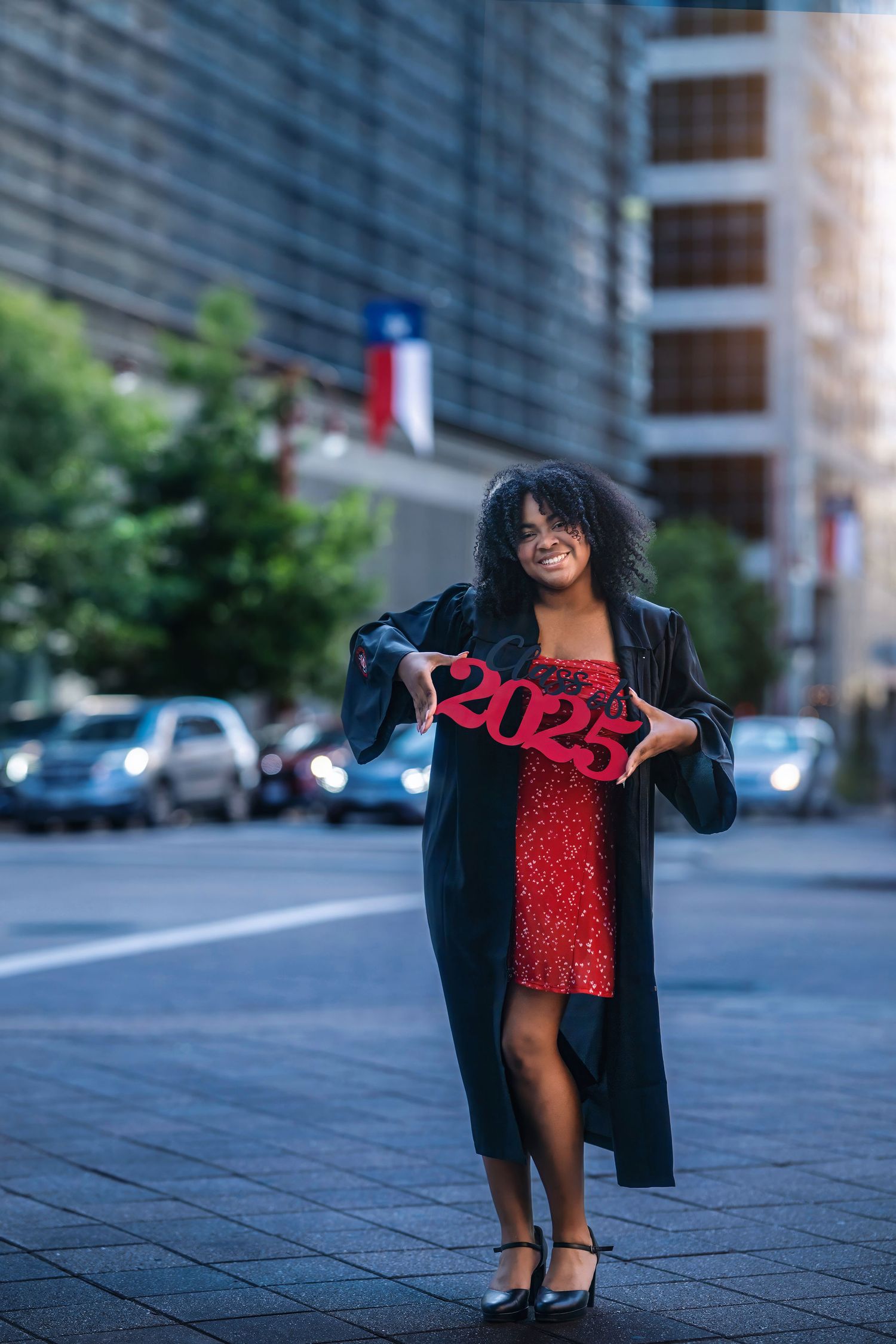 Smiling graduate in black gown and red dress holding a 2025 sign on a city street.