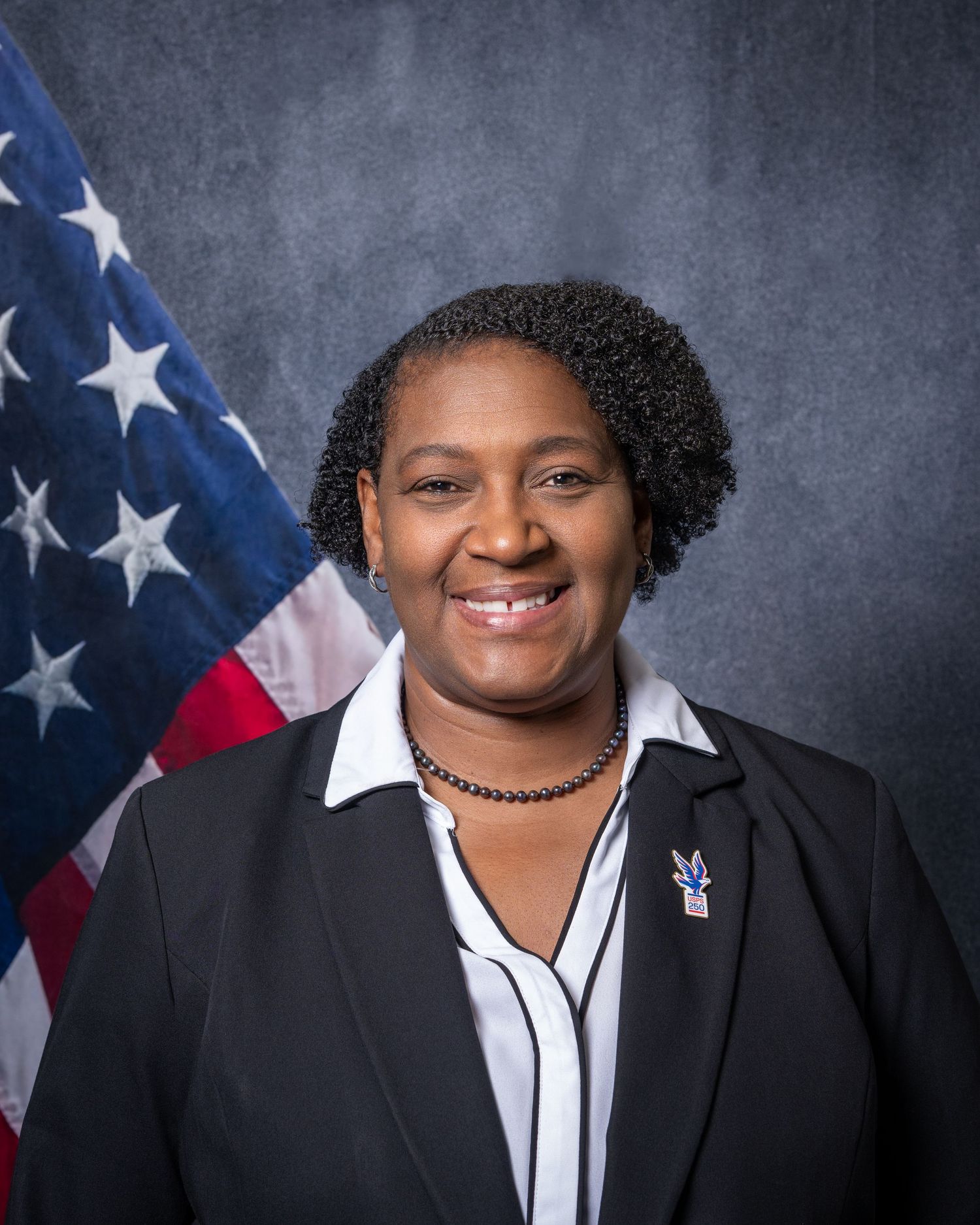 Official headshot of a woman in a black blazer smiling before an American flag backdrop.
