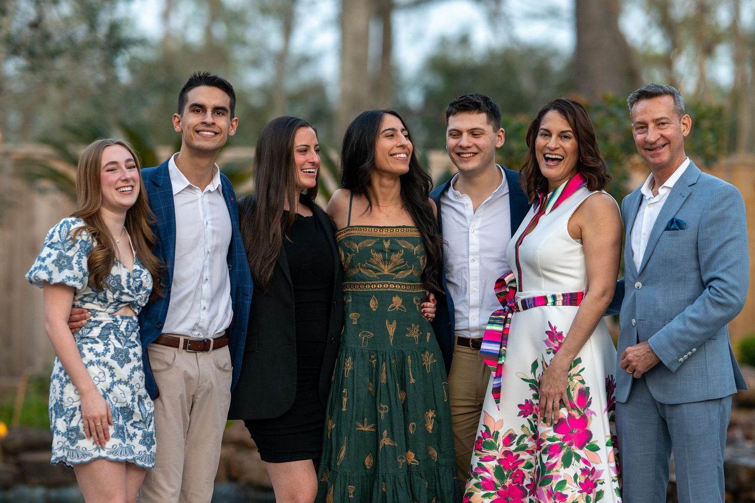 Seven smiling people dressed in formal attire posing together outdoors at a celebratory event.
