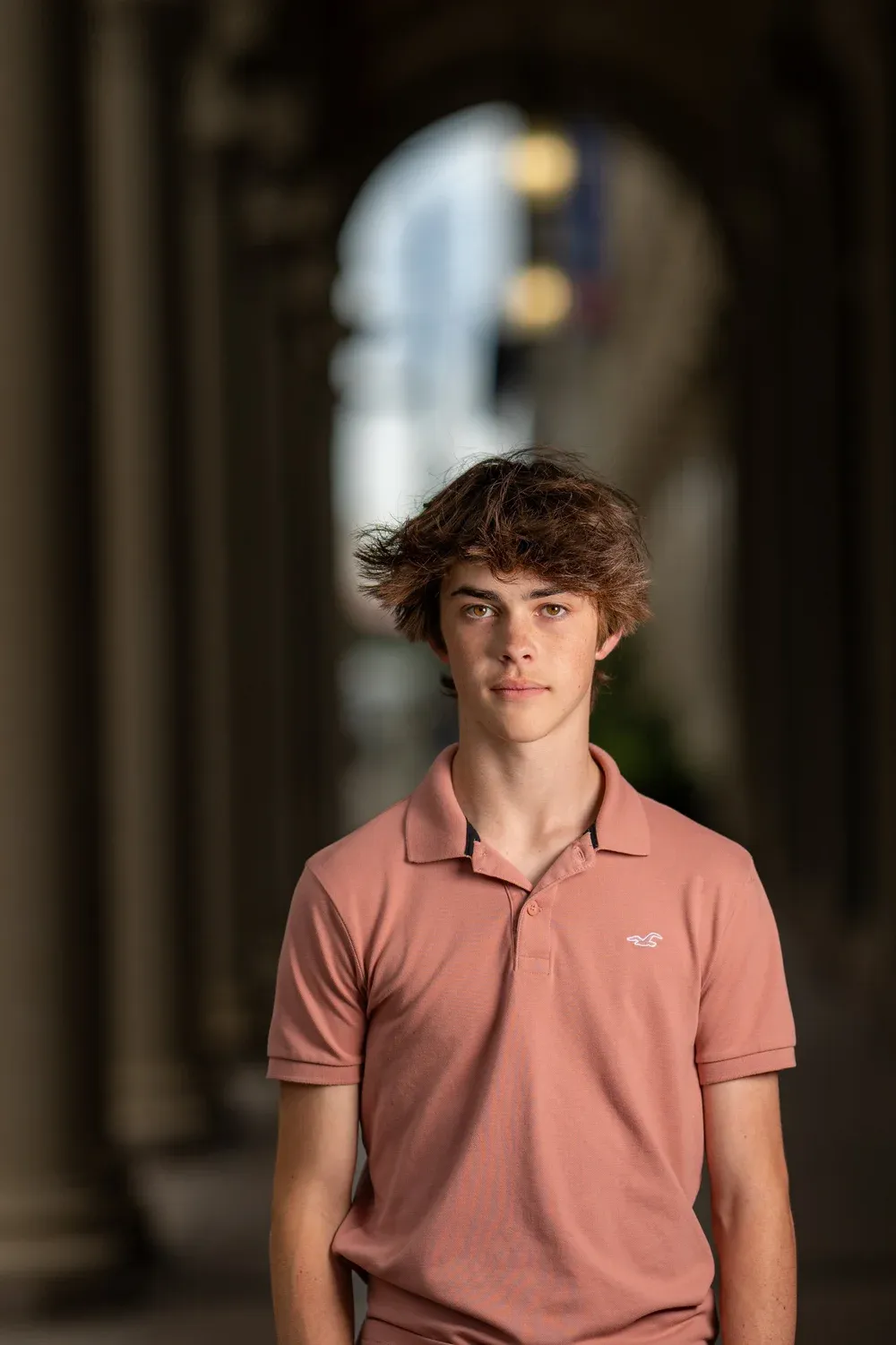 Young man with curly brown hair wearing a pink polo shirt standing under archways.