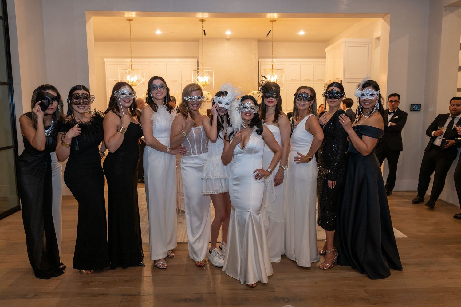 Group of women in formal black and white gowns posing together wearing decorative masquerade masks at event.