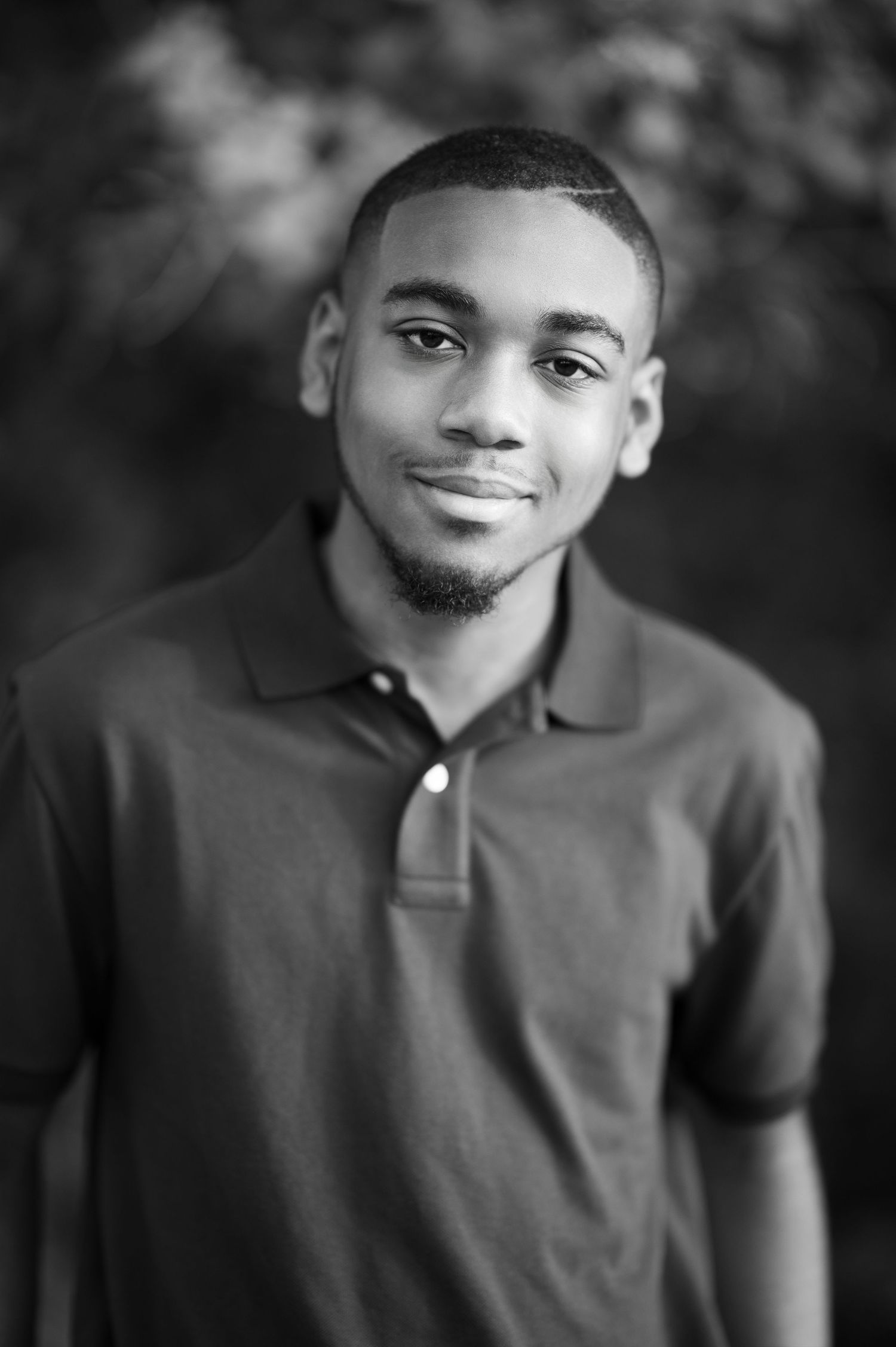 Black and white portrait of a young Black man in a polo shirt smiling outdoors.