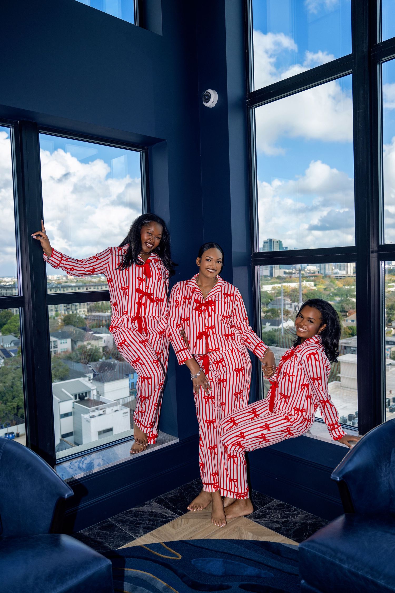 Three women in matching red and white striped pajamas posing by floor-to-ceiling windows with city view.