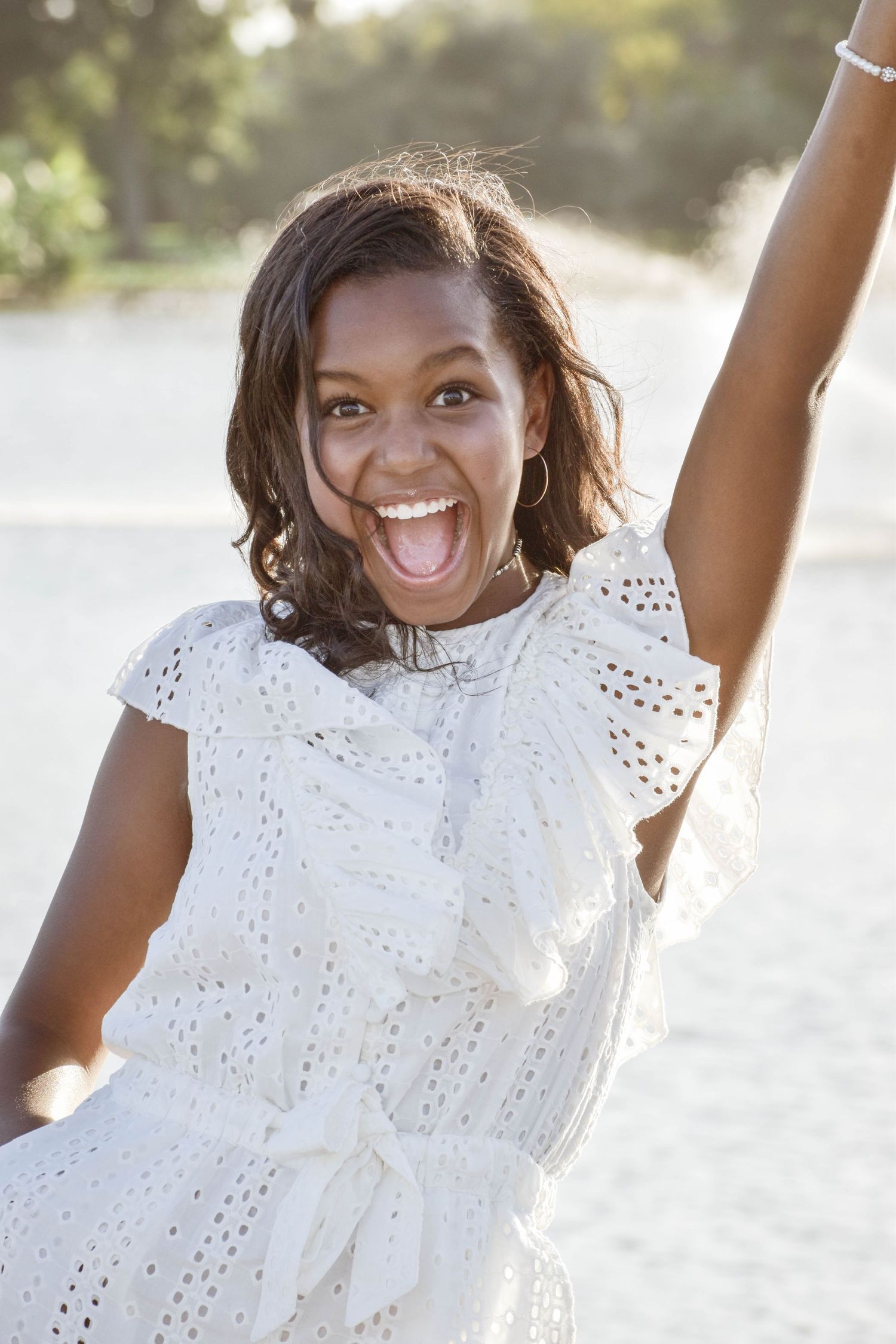 Excited young Black woman in white eyelet dress raises her arm and laughs on a sunny beach.