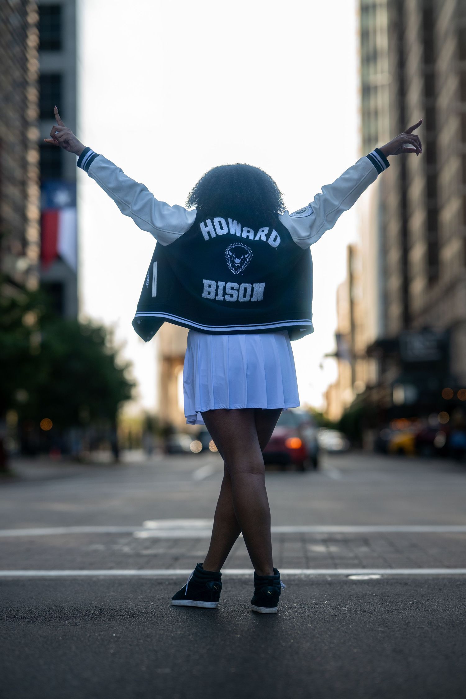 Woman in Howard Bison jacket raises arms wide while standing in the middle of a city street.
