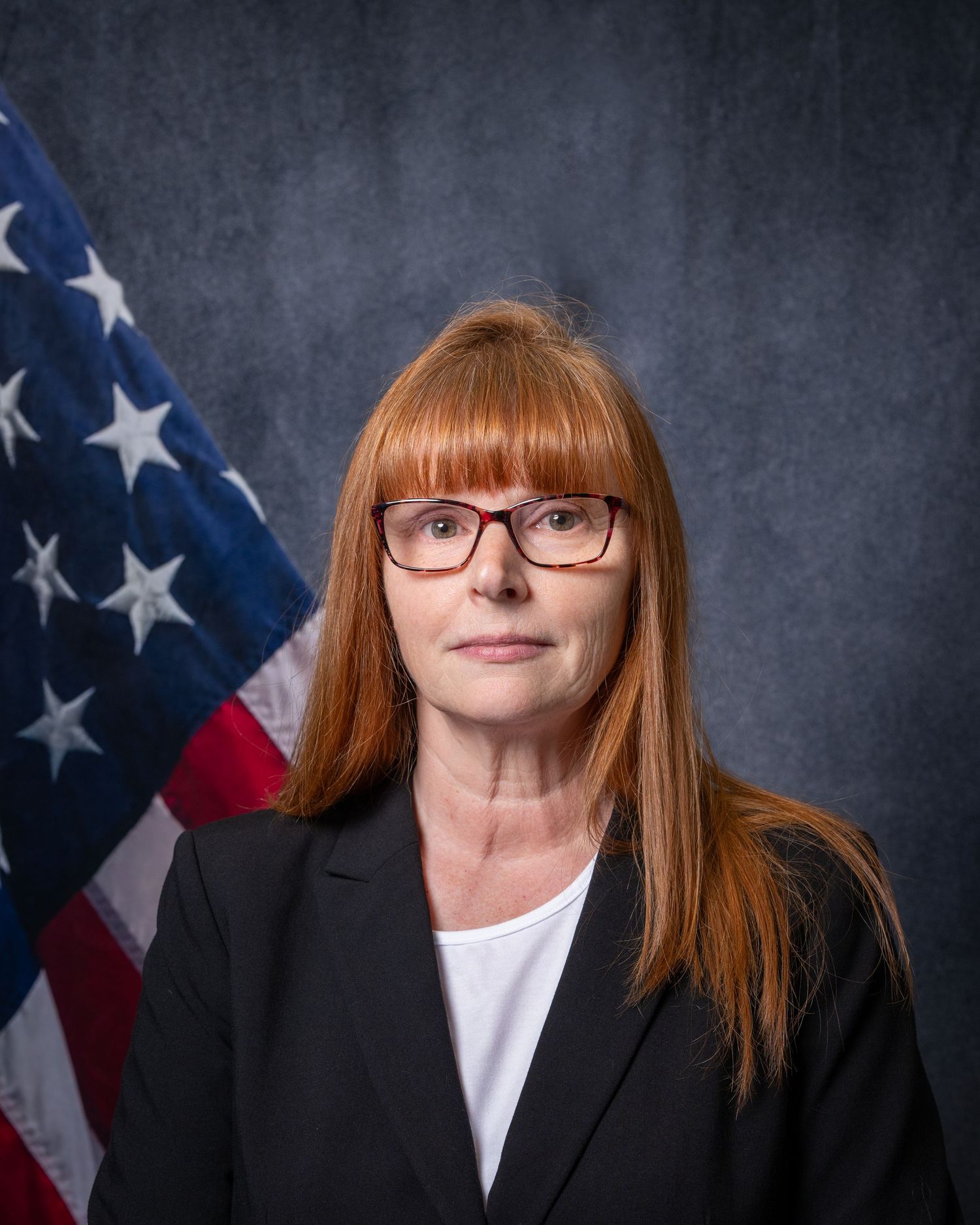 Professional headshot of a woman with red hair and glasses in a black blazer before an American flag.