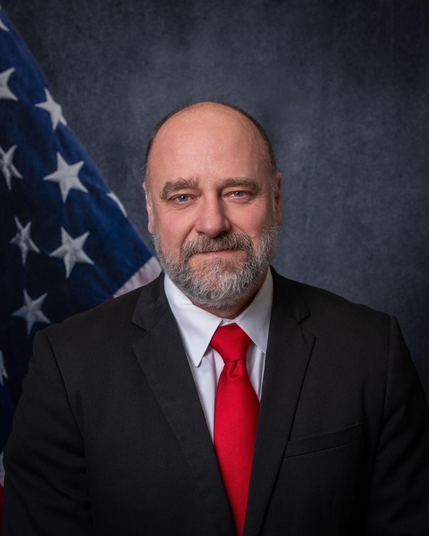 Official portrait of a bald bearded man in a black suit and red tie posing before an American flag.
