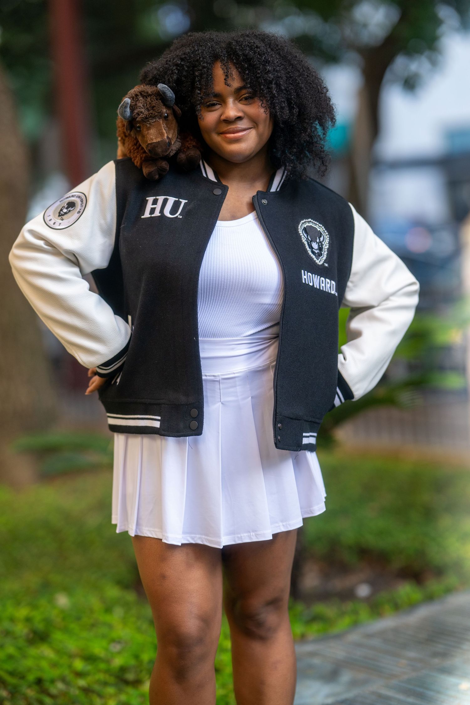 Smiling young Black woman in Howard University varsity jacket and white dress with bison plush toy.