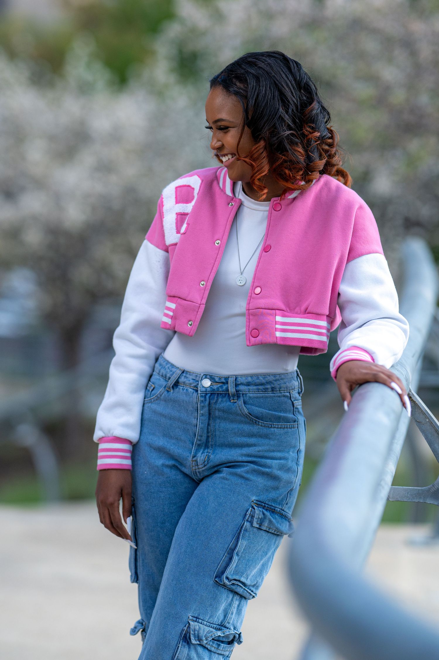 A smiling young woman in a pink varsity jacket and cargo jeans leans against a railing outdoors.