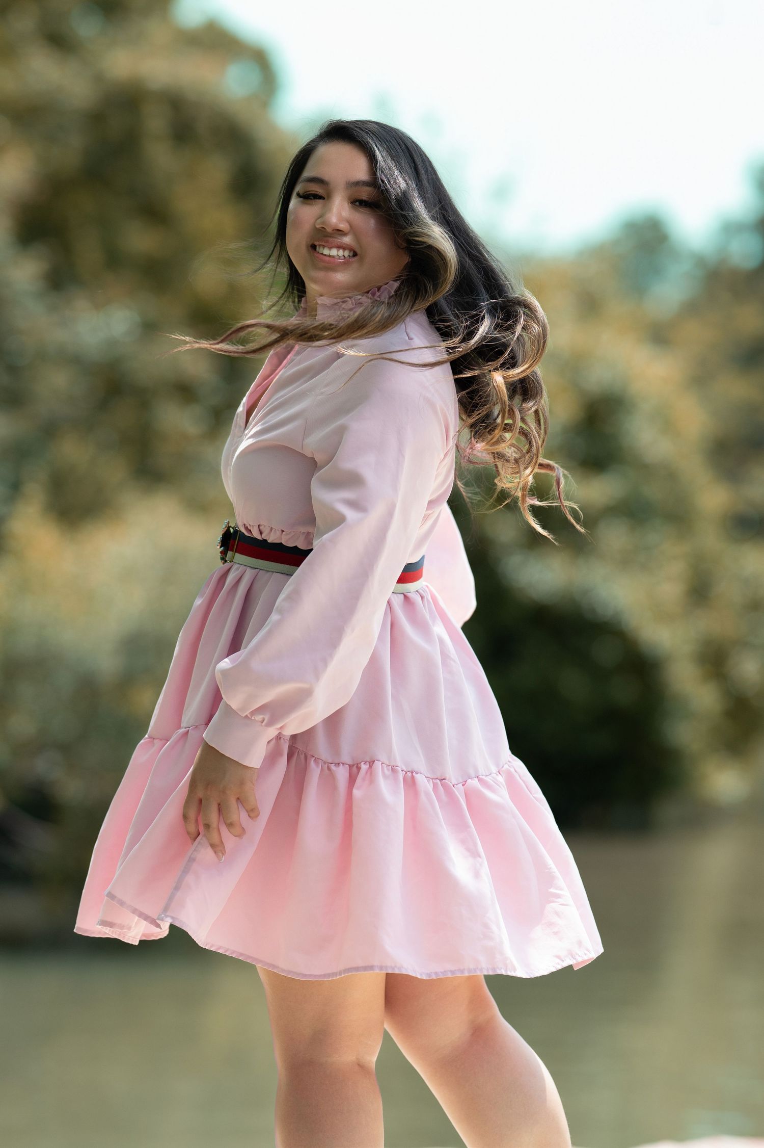 A smiling woman in a pink tiered dress twirls outdoors near a lake with trees in the background.