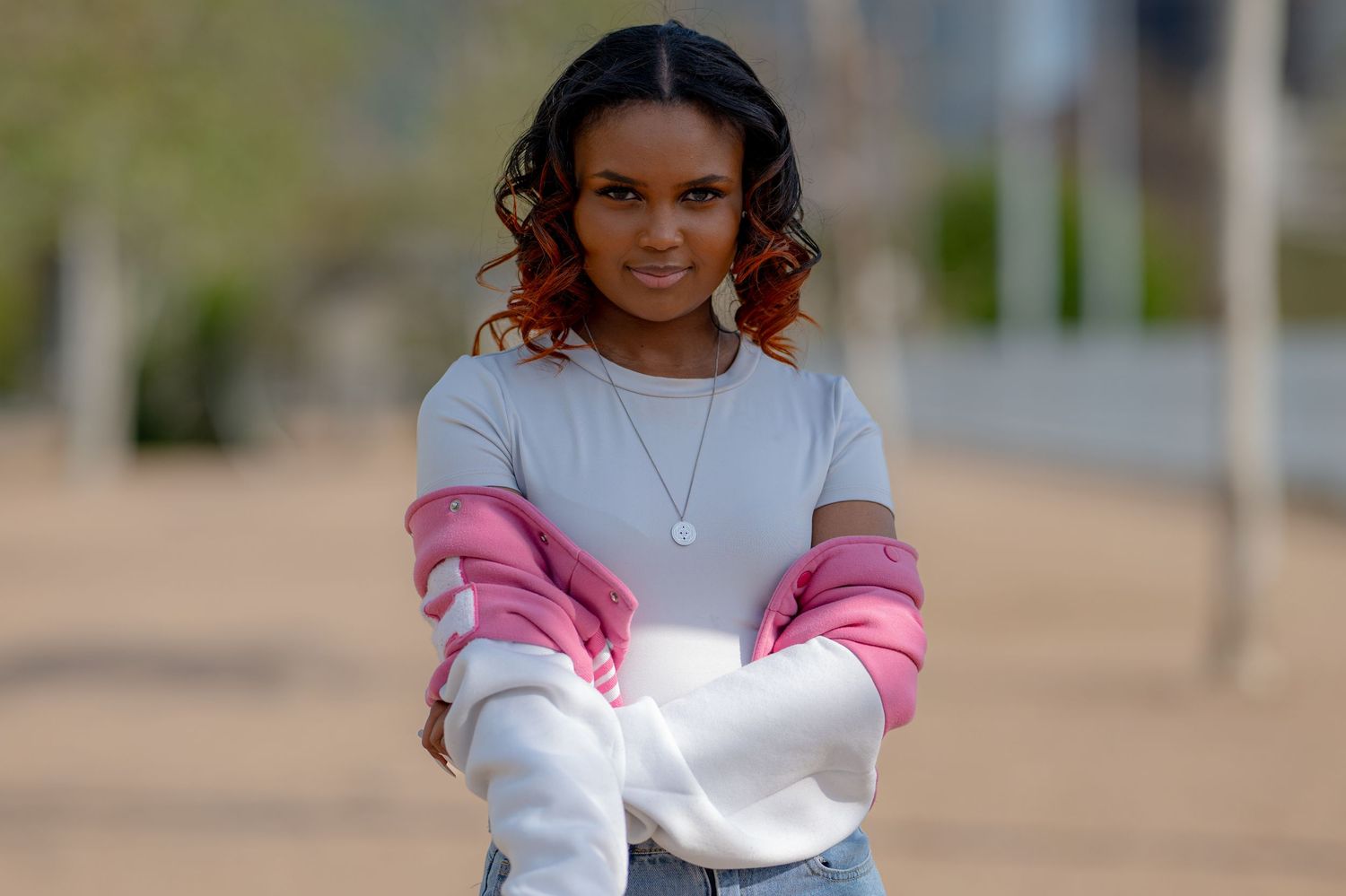 A young woman with auburn highlights stands confidently outdoors in a pink and white sweatshirt and jeans.