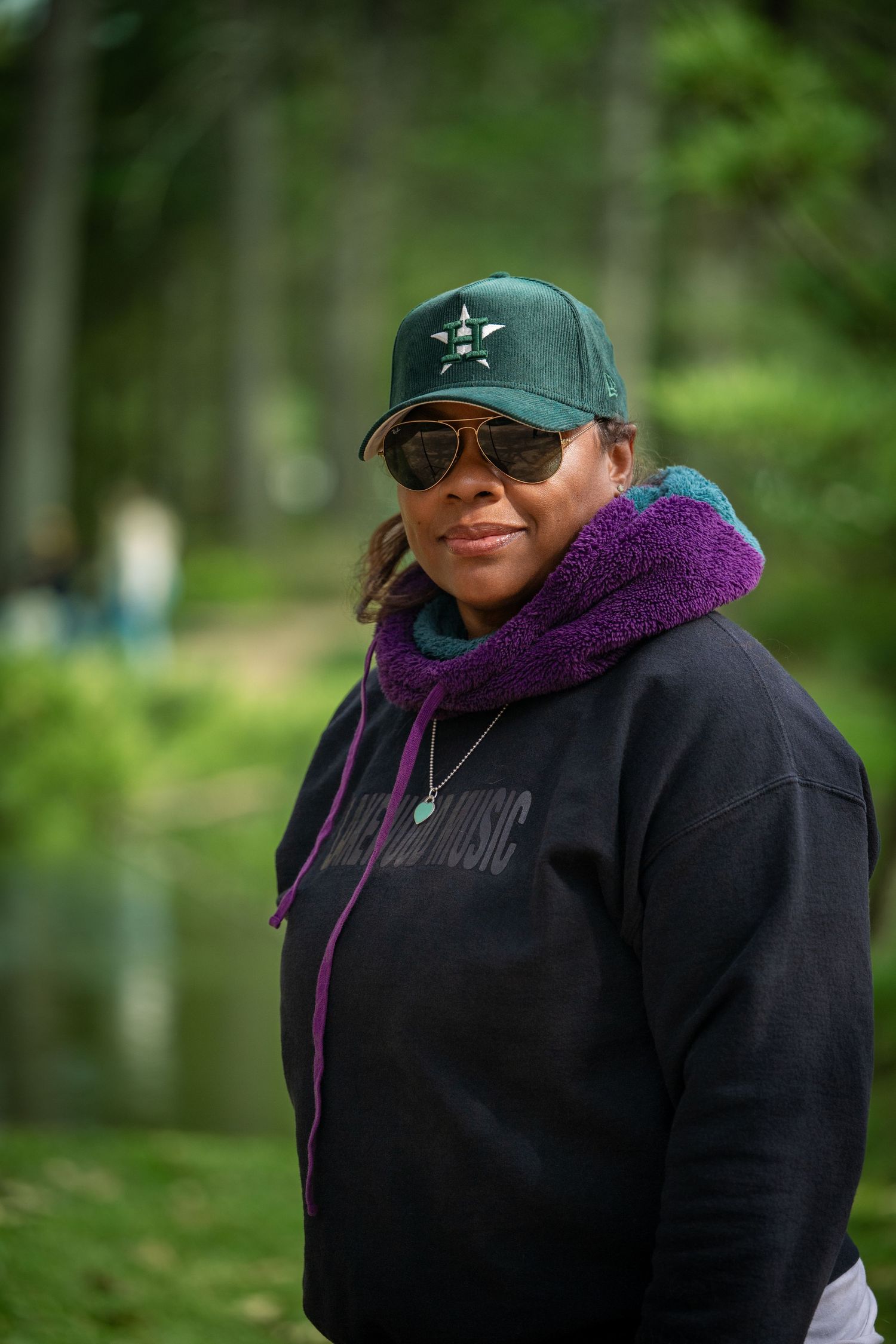 Woman in green baseball cap and aviator sunglasses wearing black hoodie stands outdoors in wooded area.