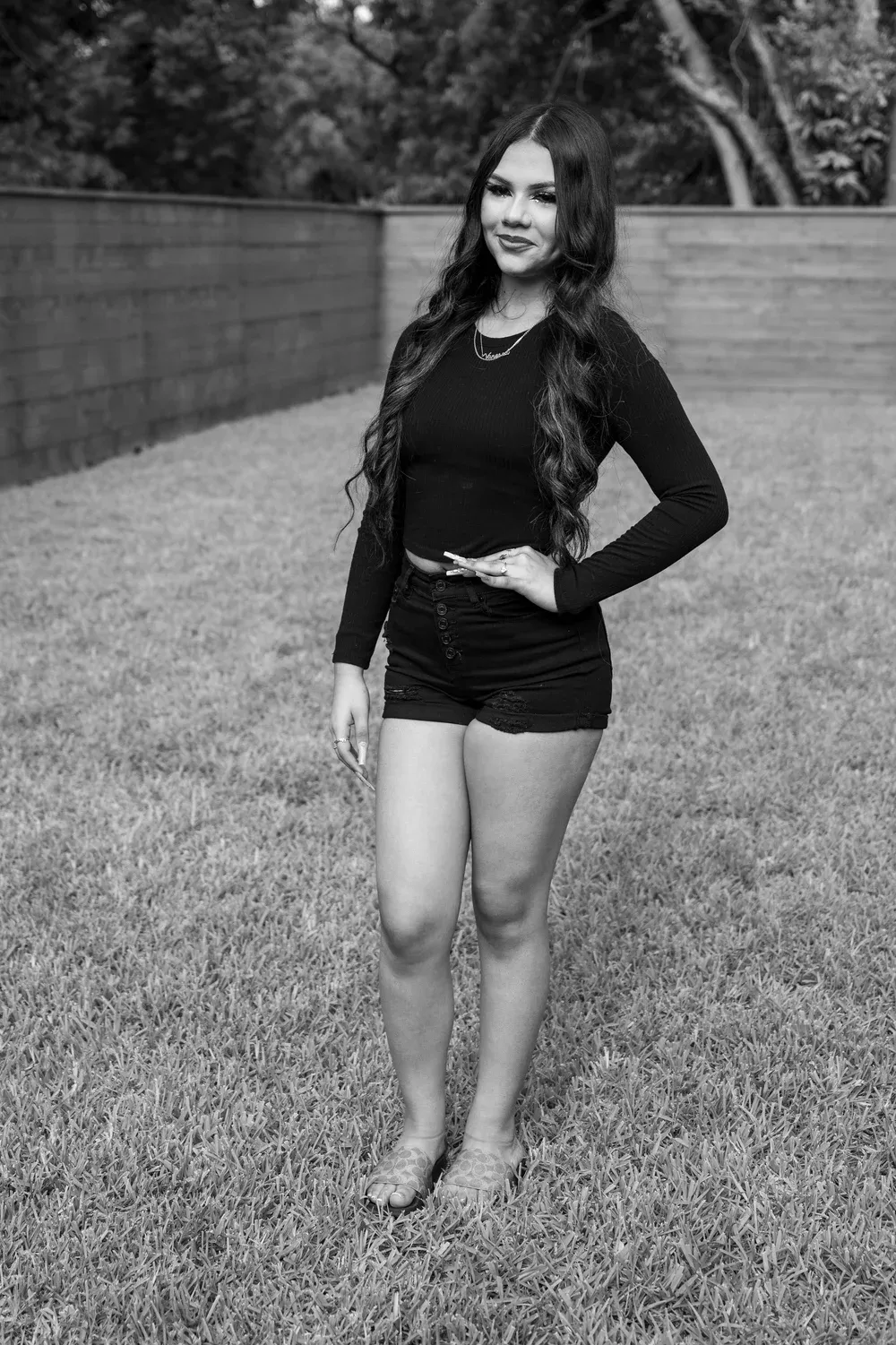 Young woman with long wavy hair in black outfit standing on green lawn in black and white portrait photo.