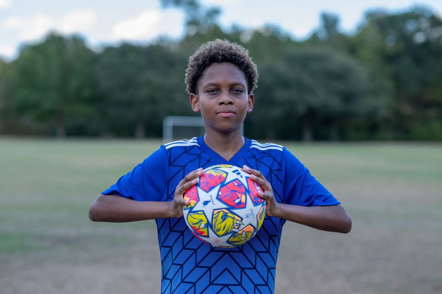 Young boy in a blue soccer jersey holding a UEFA Champions League ball on a field.