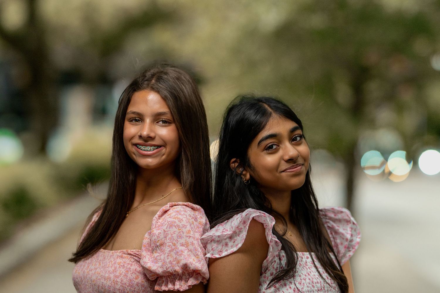 Two teenage girls in pink floral dresses smiling back-to-back on an outdoor path.