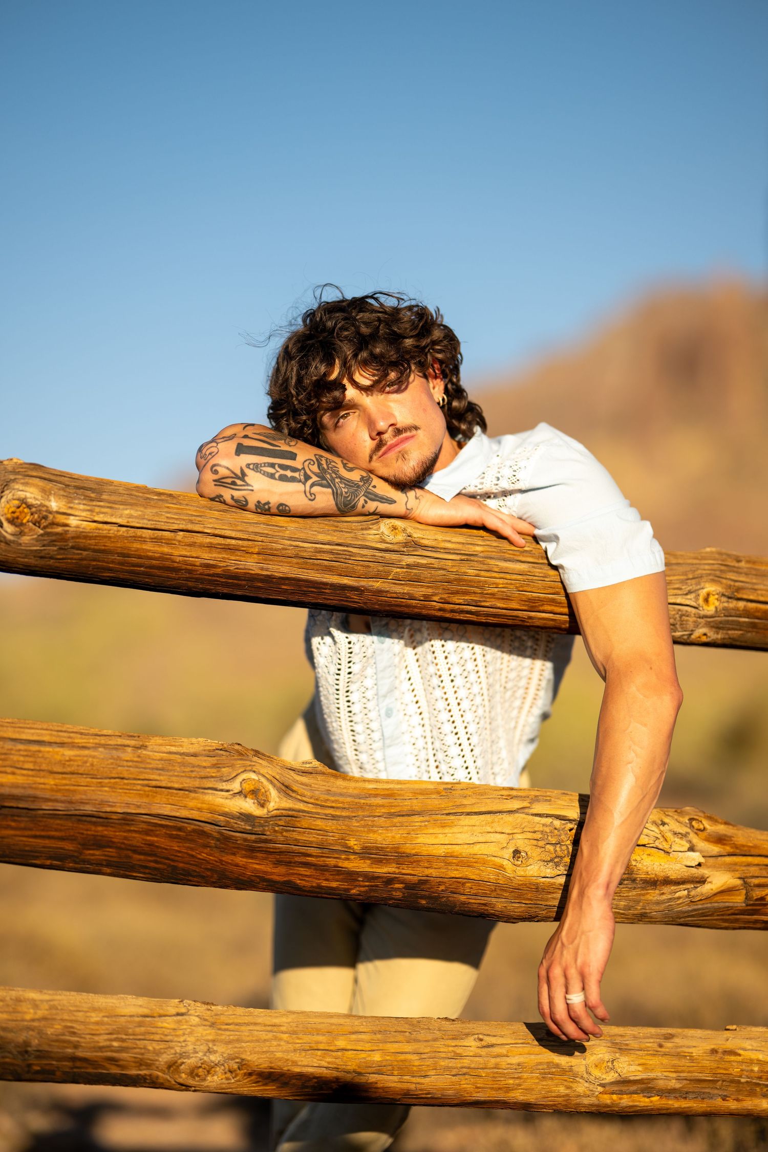 Tattooed young man with curly hair leaning on rustic wooden fence in desert landscape under blue sky.