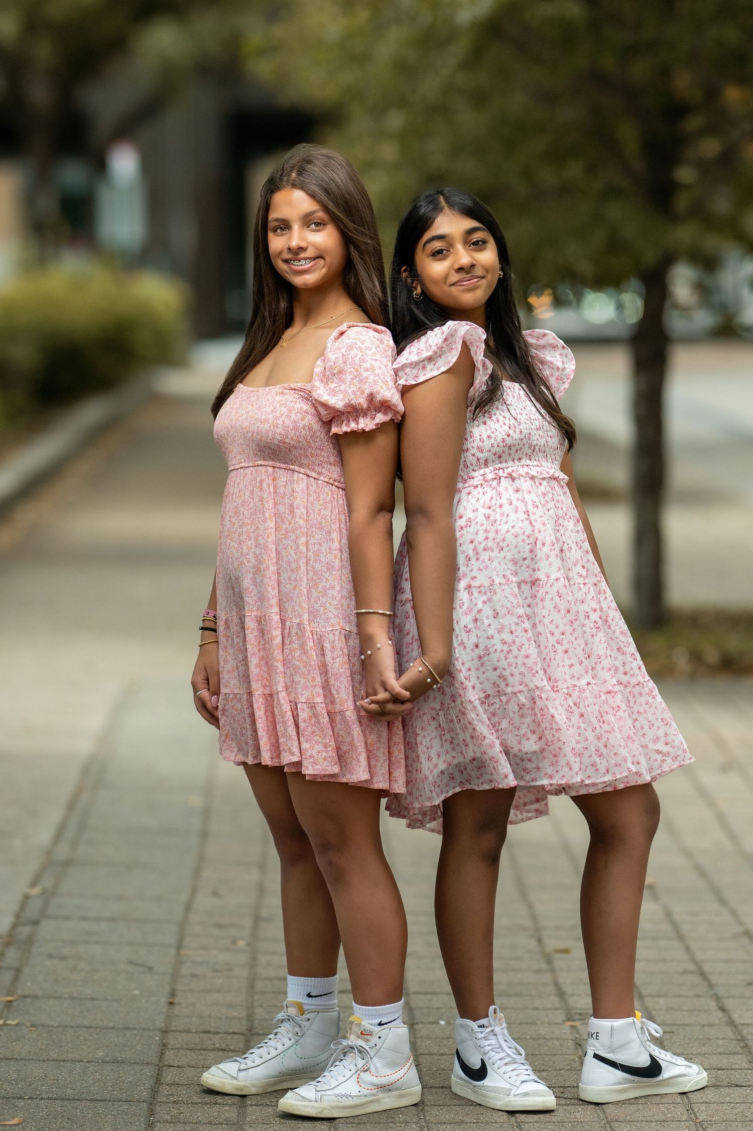 Two teenage girls in matching pink floral dresses holding hands while posing outdoors on a sidewalk.