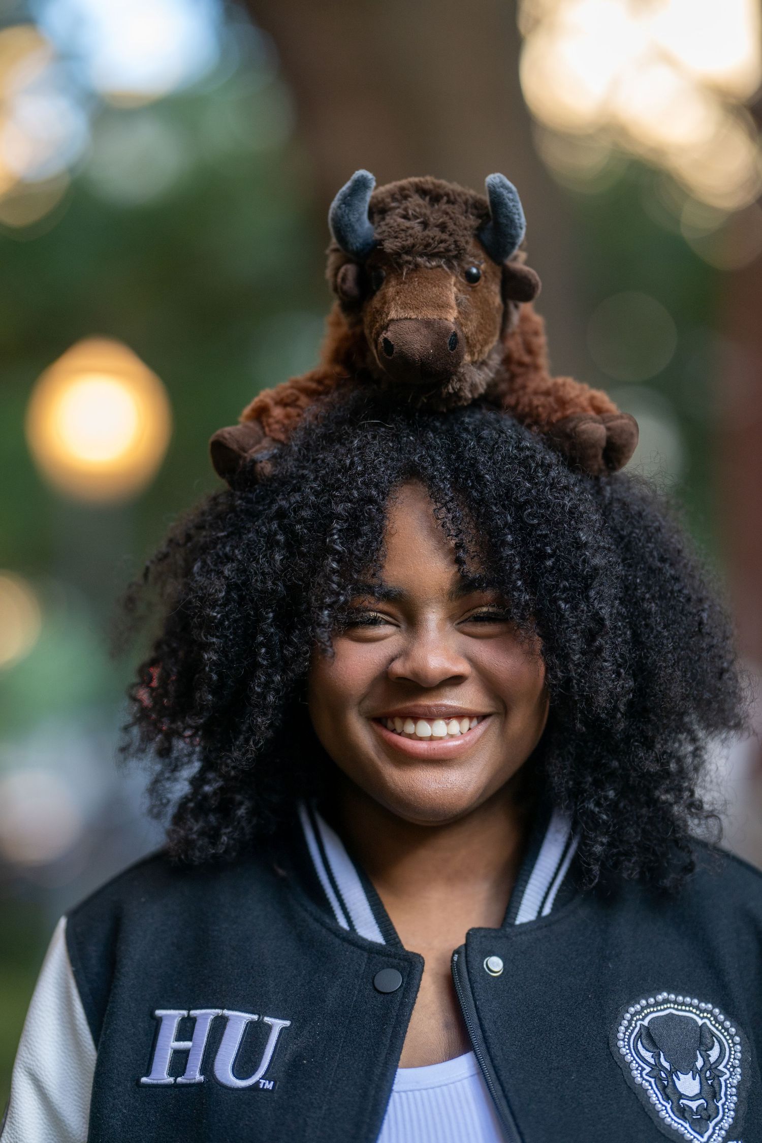 Joyful young Black woman smiling with a bison stuffed animal on her head wearing an HU jacket.