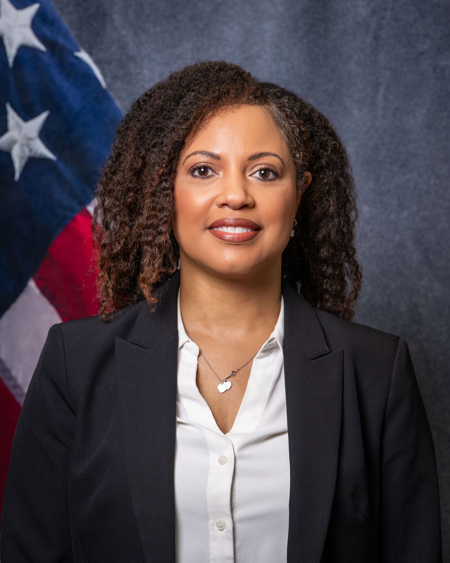 Professional portrait of a woman in a black blazer posing before an American flag backdrop.