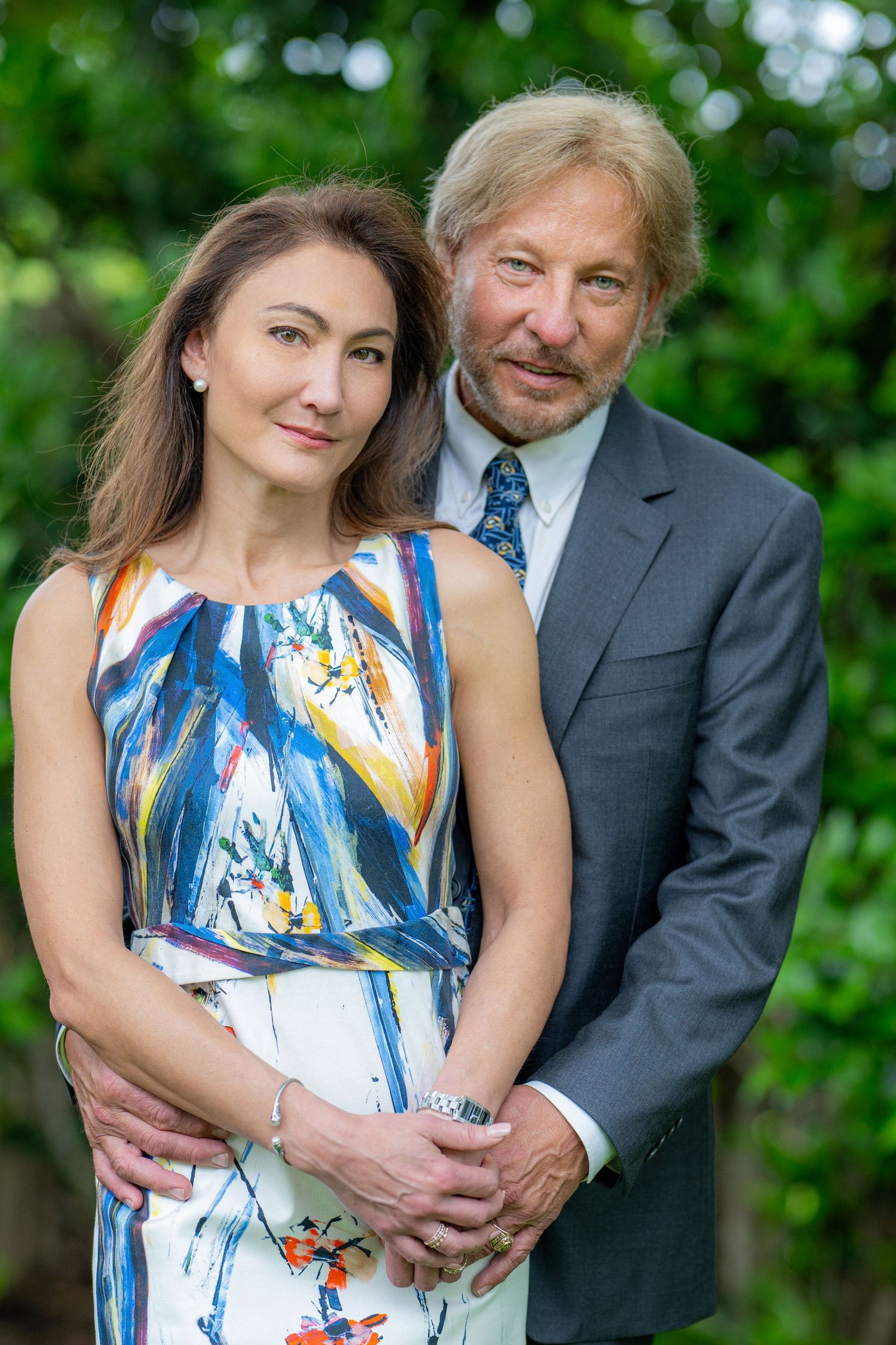 An elegant couple poses together outdoors, the woman in a colorful dress and the man in a gray suit.