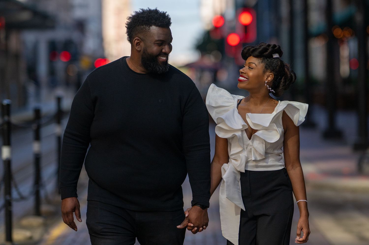 A couple walks hand in hand down a city street, smiling at each other affectionately.