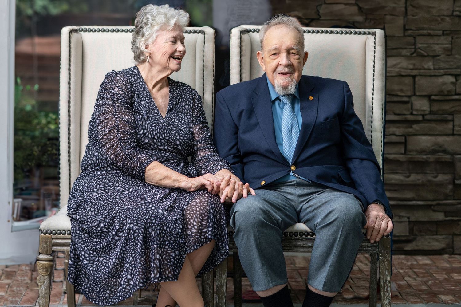 An elderly couple sits in elegant chairs, holding hands and laughing joyfully together.