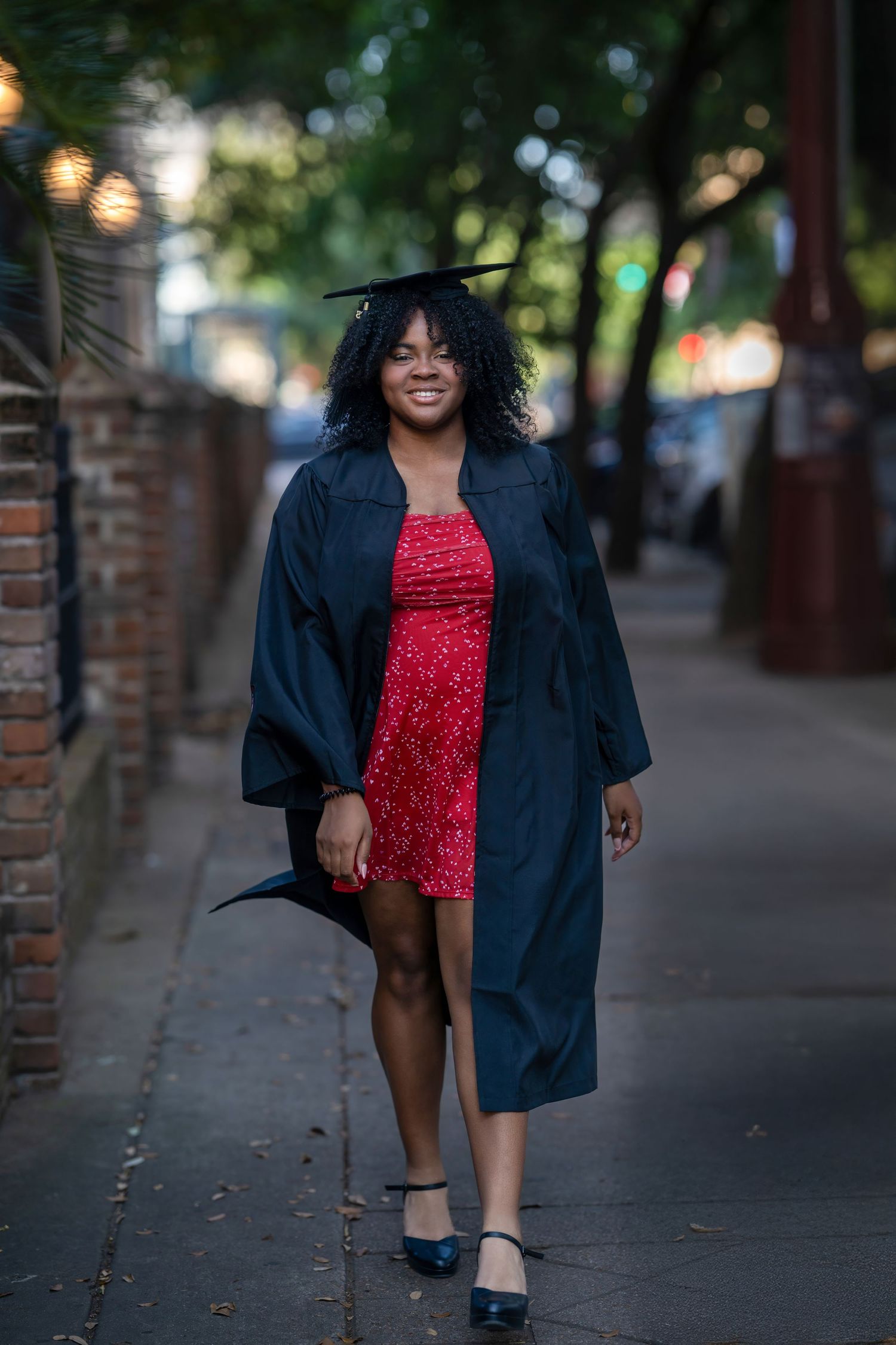 A smiling woman in a red dress and black graduation cap and gown walks confidently down a tree-lined sidewalk.