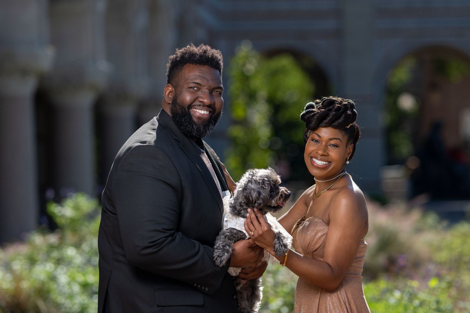 A well-dressed couple smiles while holding an adorable small dog in an outdoor setting.