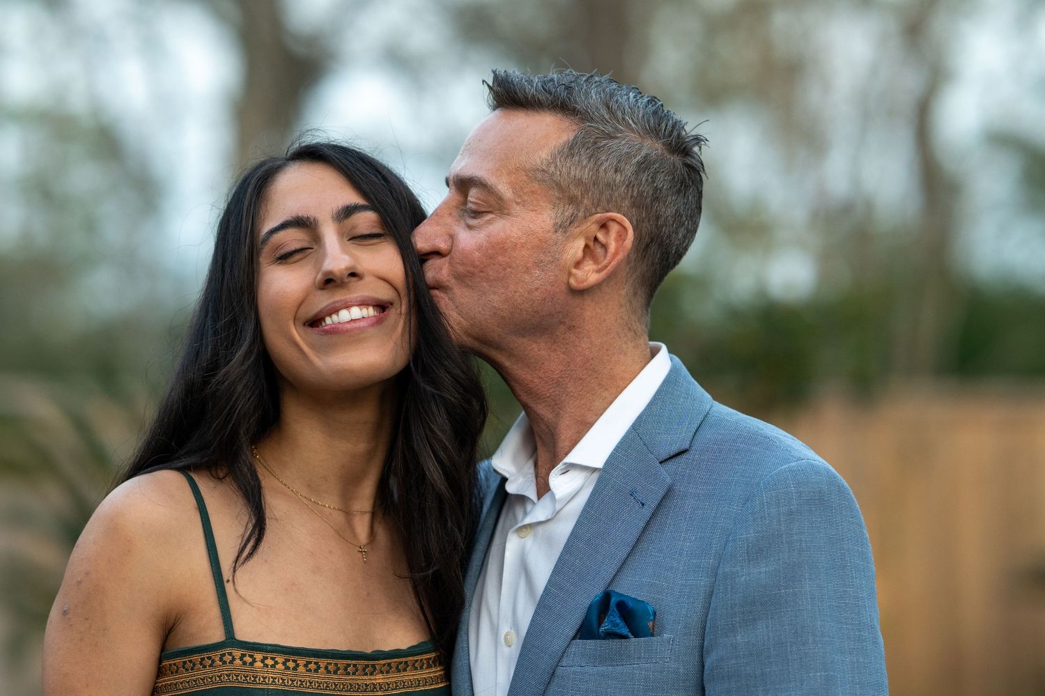 A man in a blue blazer kisses a smiling woman on the cheek outdoors in a romantic moment.