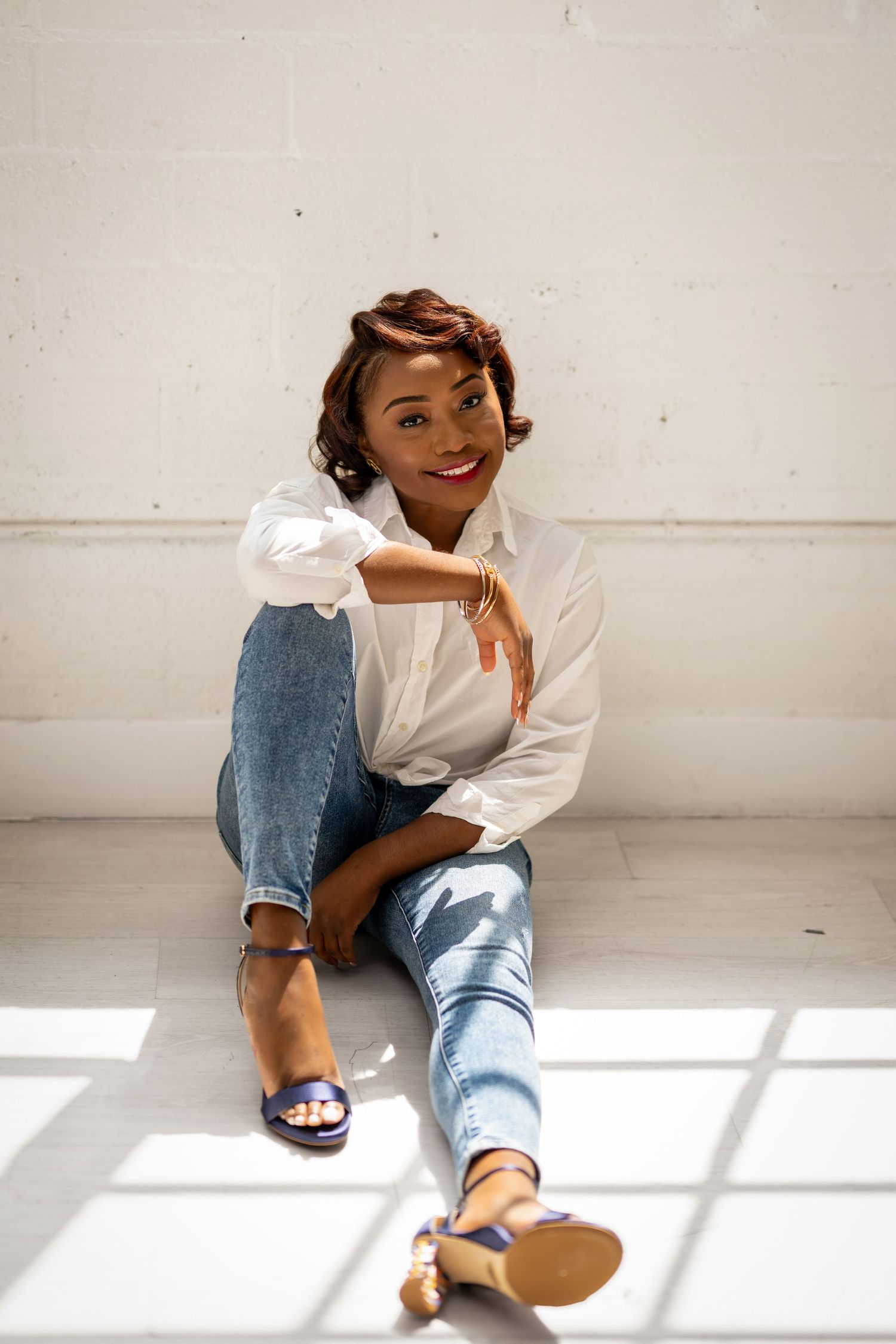 Smiling woman in white shirt and jeans sitting on floor against white wall, wearing blue heeled sandals.