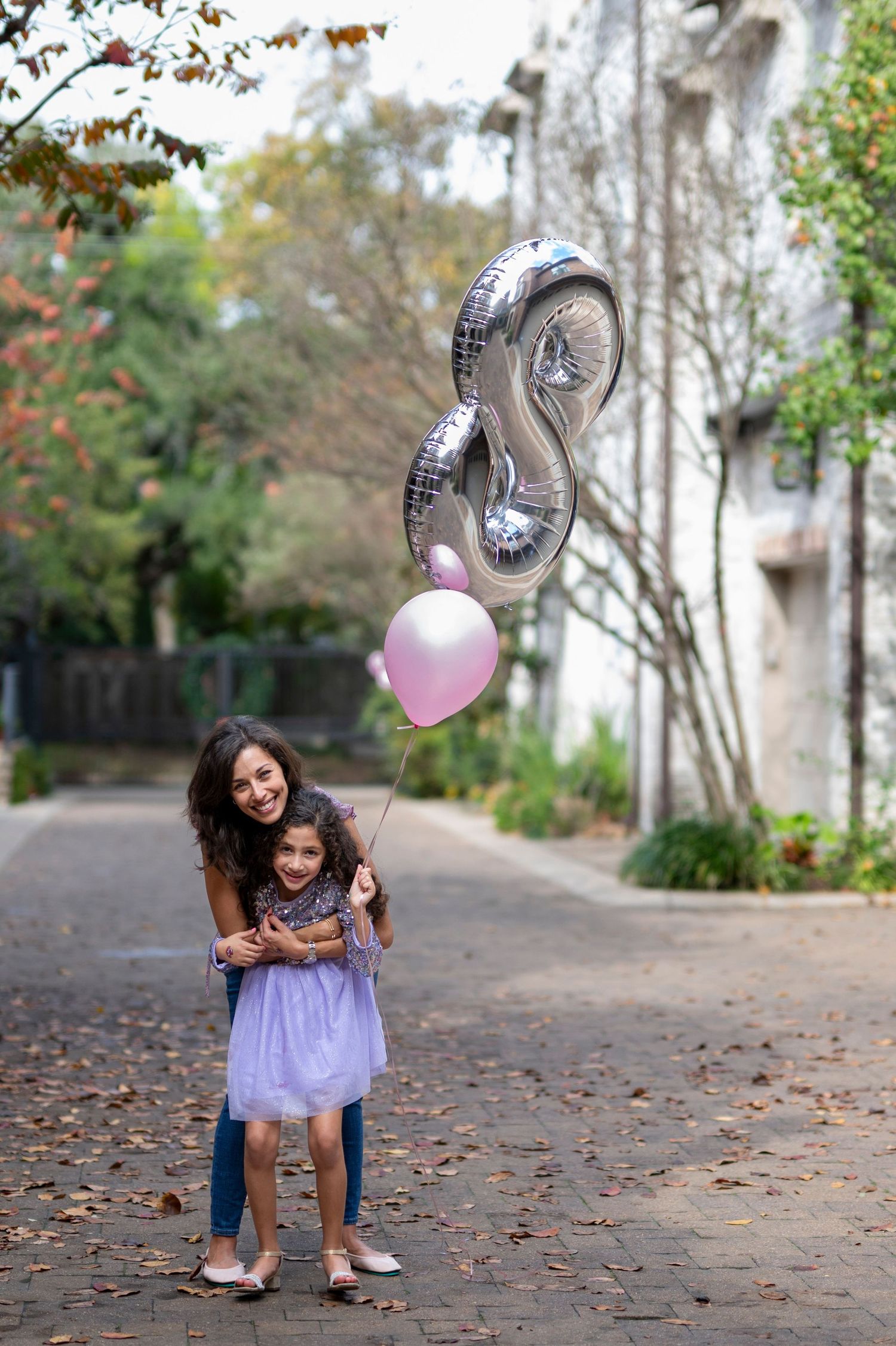 Smiling mother embracing young daughter holding pink and silver number 8 balloons on tree-lined path.
