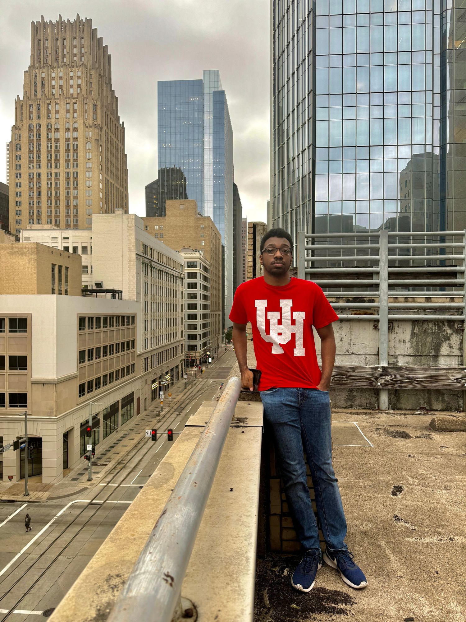 Young man in red University of Houston t-shirt posing on rooftop with downtown city skyline behind him.