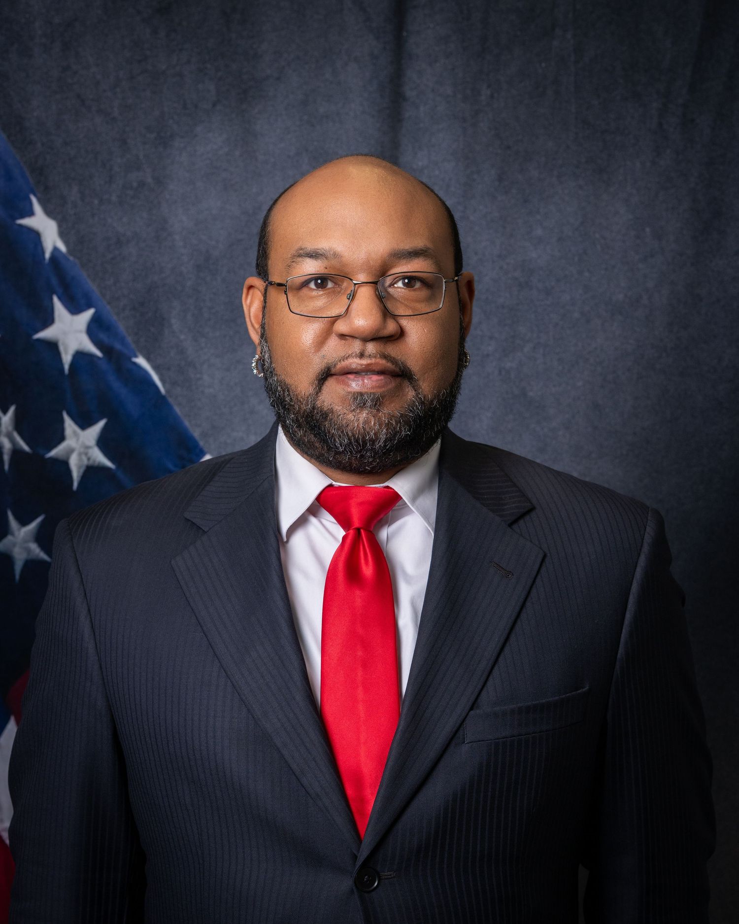 Professional headshot of a bearded man with glasses in a pinstripe suit and red tie before an American flag.