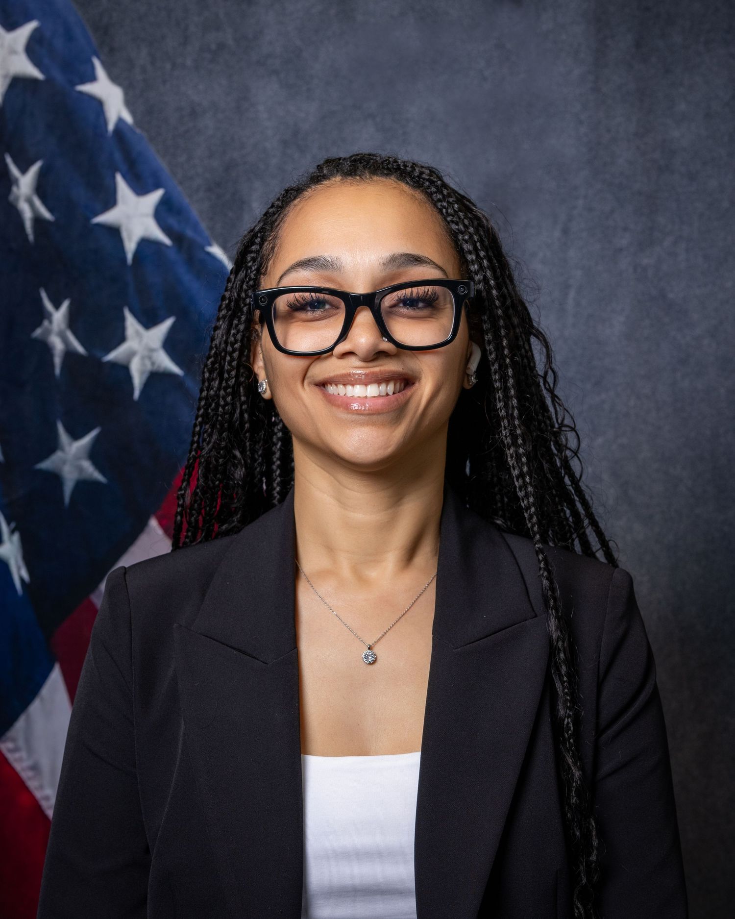Professional portrait of a young woman with braids and glasses smiling before an American flag.